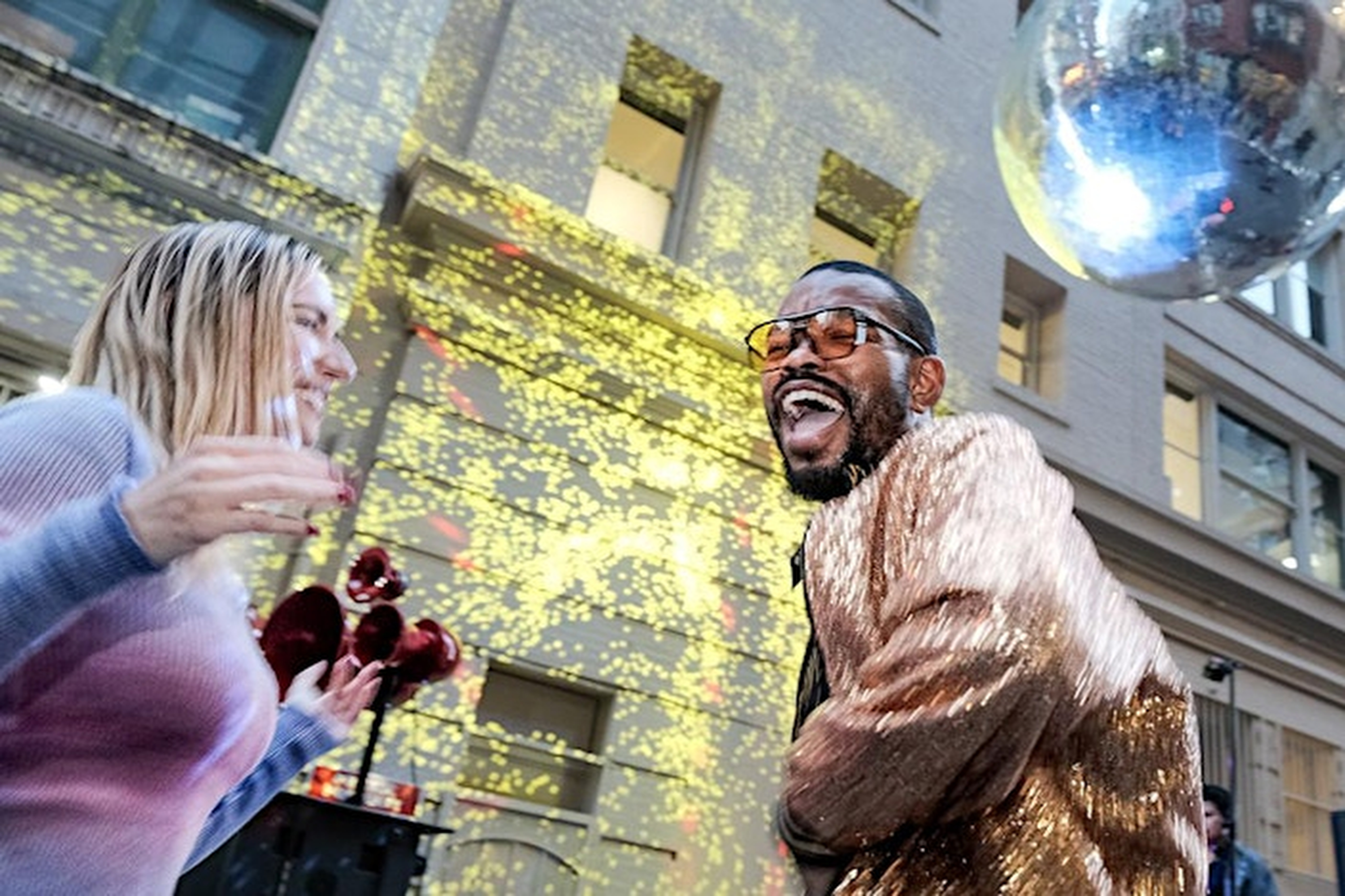 Two people are joyfully dancing outdoors. One wears a sparkly gold jacket, and the other a light sweater. A disco ball reflects light on a nearby building.