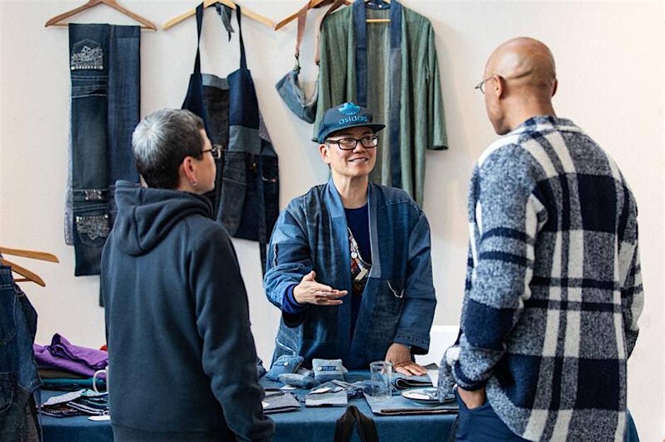 Three people are discussing in front of a table with fabric items. One wears a cap and gestures while speaking. Garments hang on the wall behind them.