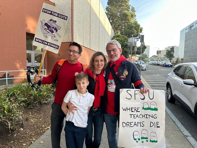 A group of four people, including a child, stand together on a sidewalk holding protest signs about SFSU. They appear cheerful and united in a public area.