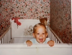 A baby with blonde hair is smiling, standing in a white crib. The room has floral wallpaper with red and pink flowers and a brown teddy bear is in the crib.