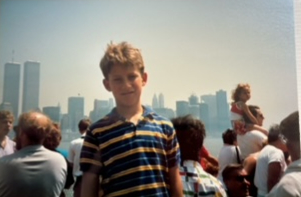 A boy in a striped shirt stands in a crowded area with a hazy city skyline in the background, featuring two tall towers.