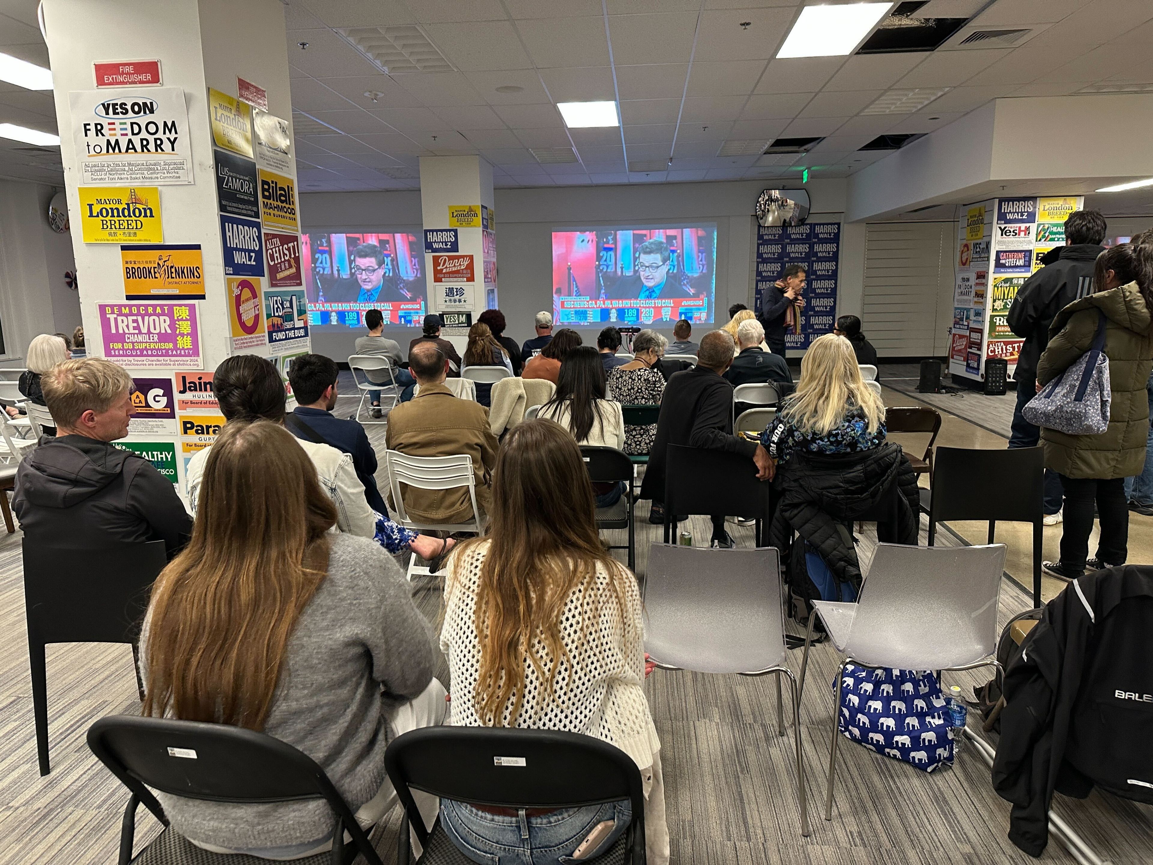 A group of people is seated in a room watching a TV screen displaying a man speaking, possibly at a political event. Campaign signs cover the walls.