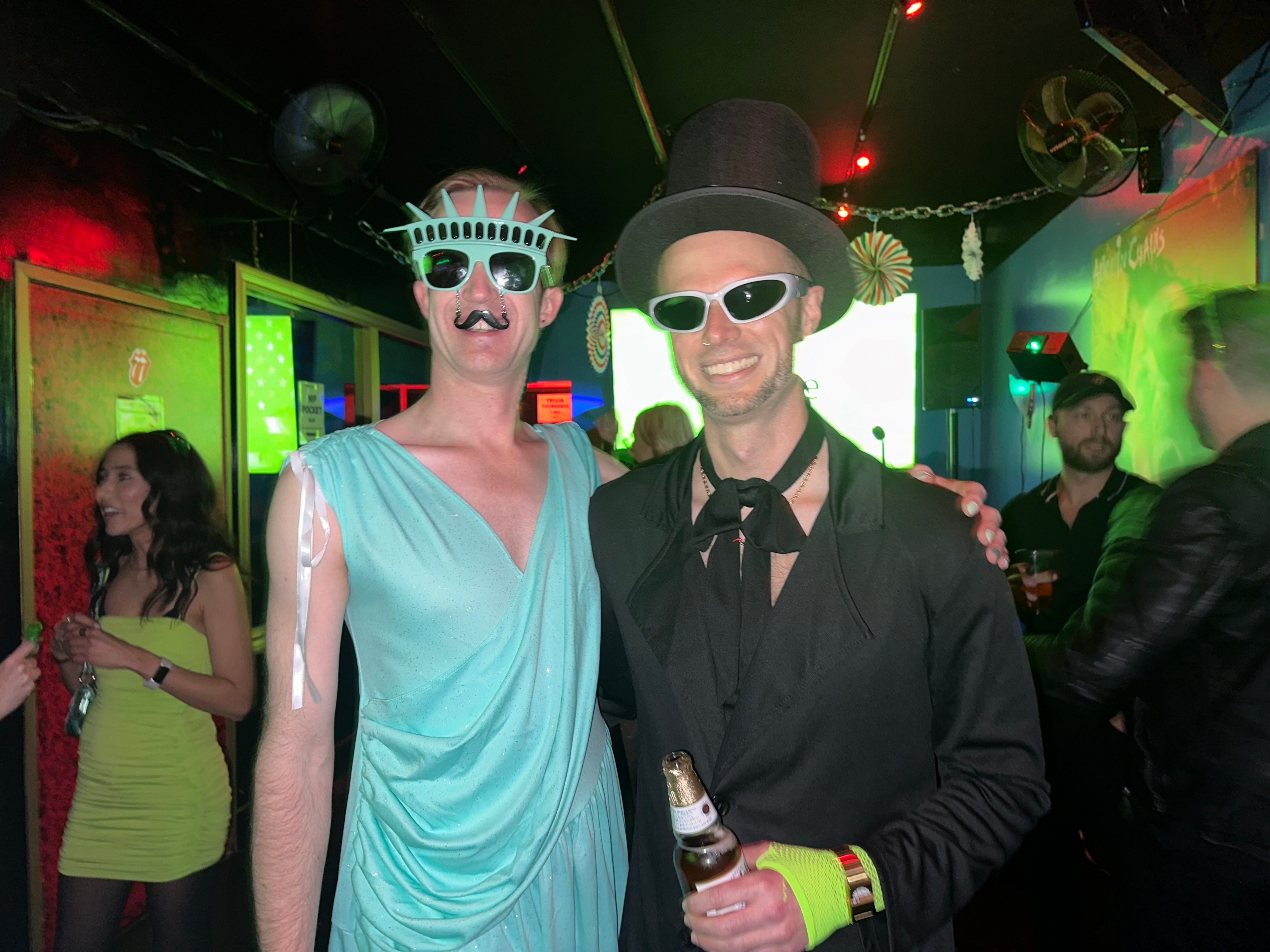 Two people in costumes pose together; one wears a Statue of Liberty outfit with sunglasses and a mustache, the other in a black suit with a top hat and sunglasses.