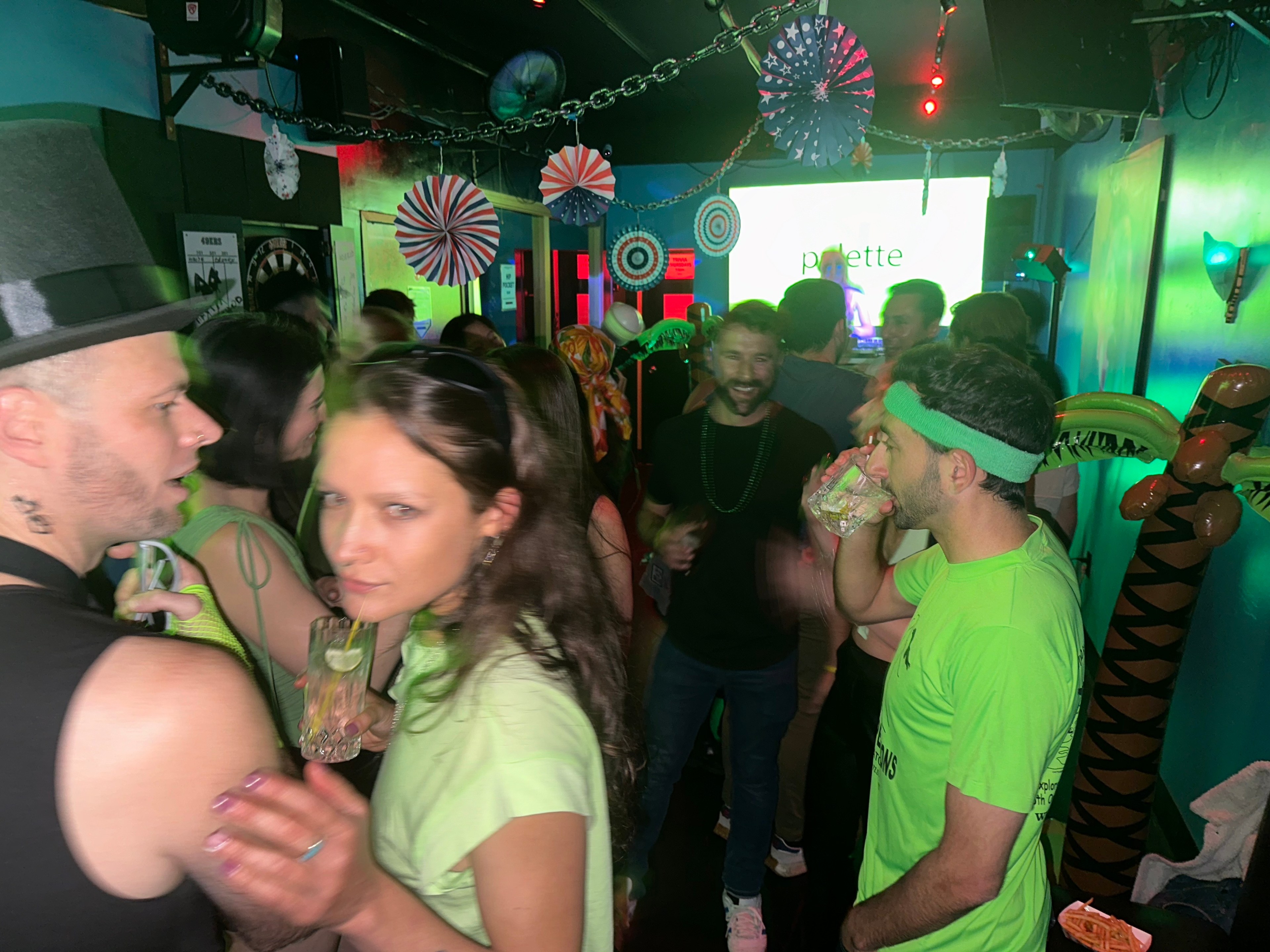 A lively party scene with people socializing and dancing in a dimly lit room. Bright green lighting, festive decor, and a man in a hat sipping a drink.