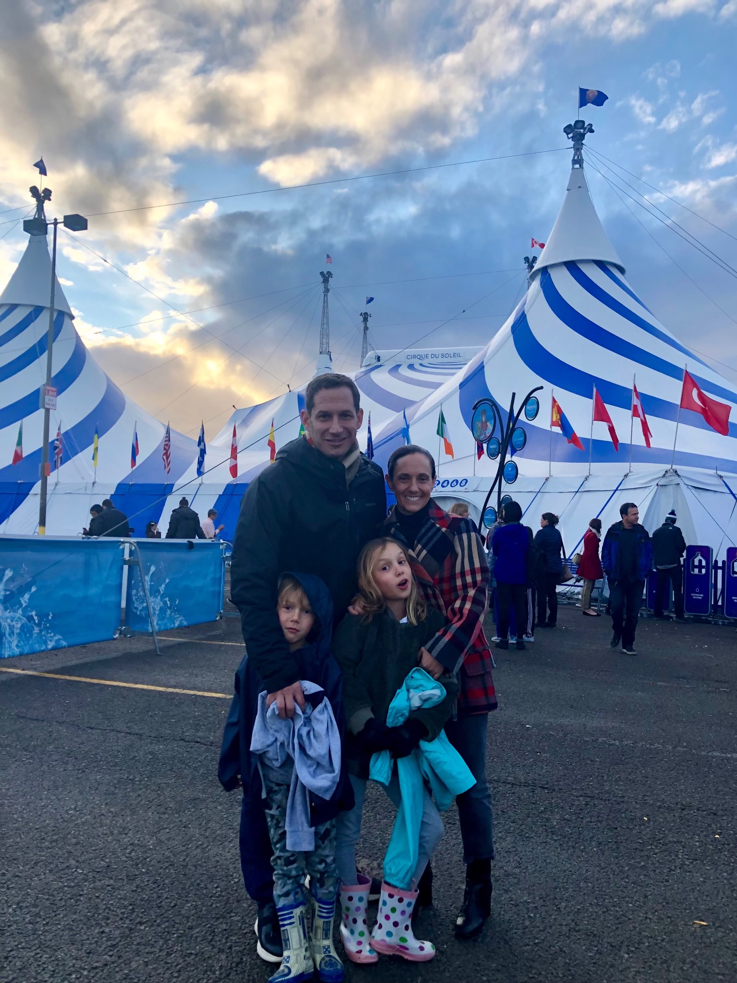 A family of four stands smiling outside a large circus tent with blue and white stripes. The sky is cloudy, and several colorful flags are visible.