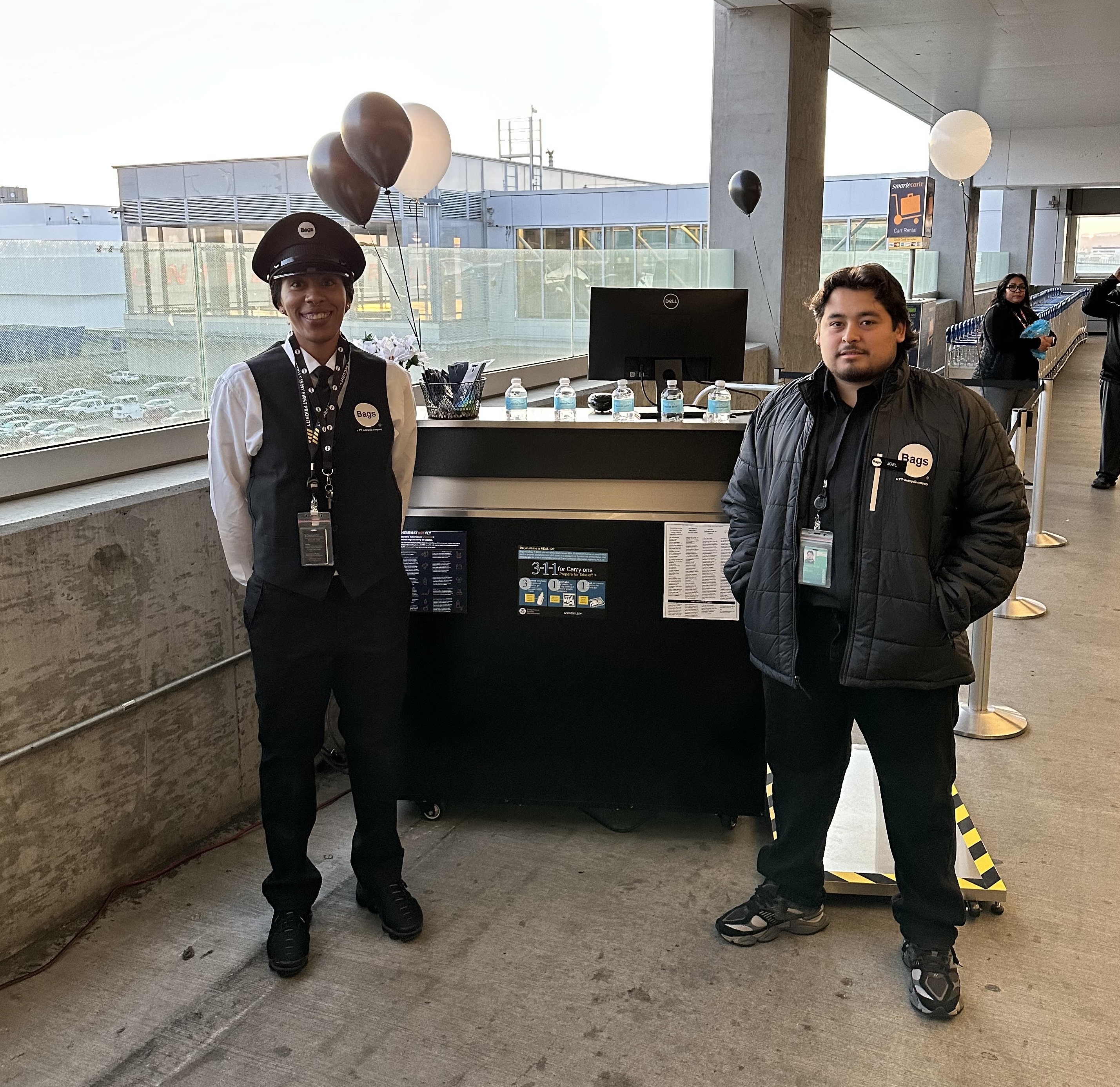 Two people in uniform stand by a counter with balloons. One wears a cap and vest, the other a jacket. Behind them is a building with large windows.