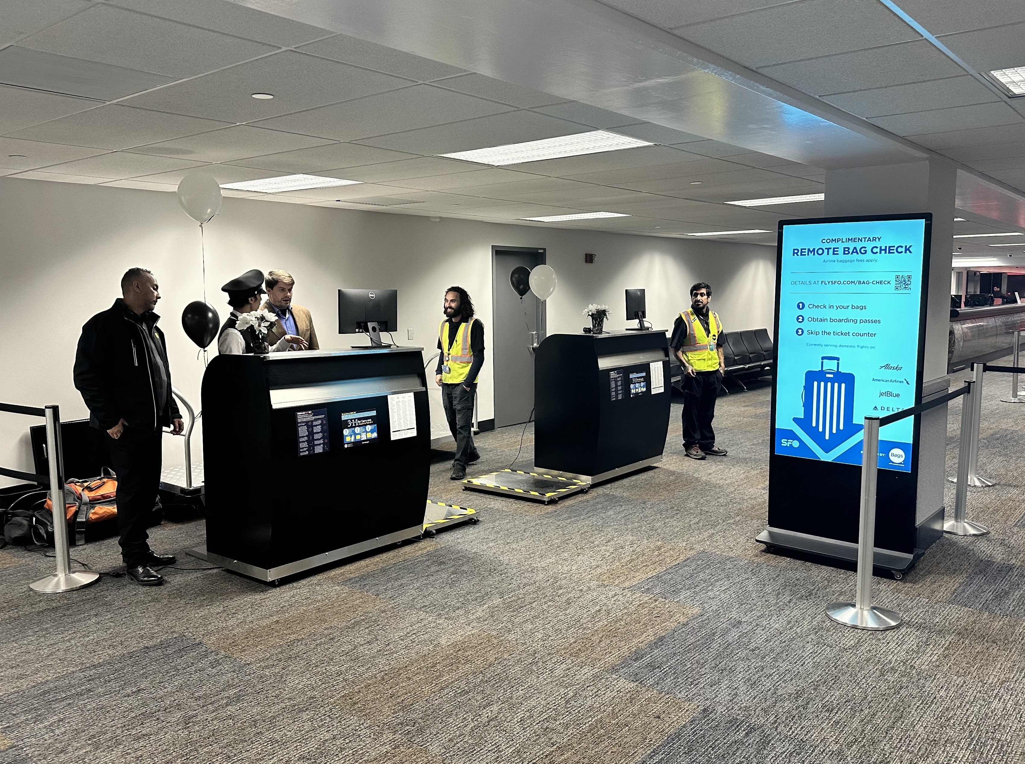 An airport area for complimentary remote bag check with two counters, staff in reflective vests, a digital sign listing airlines, and balloons as decoration.