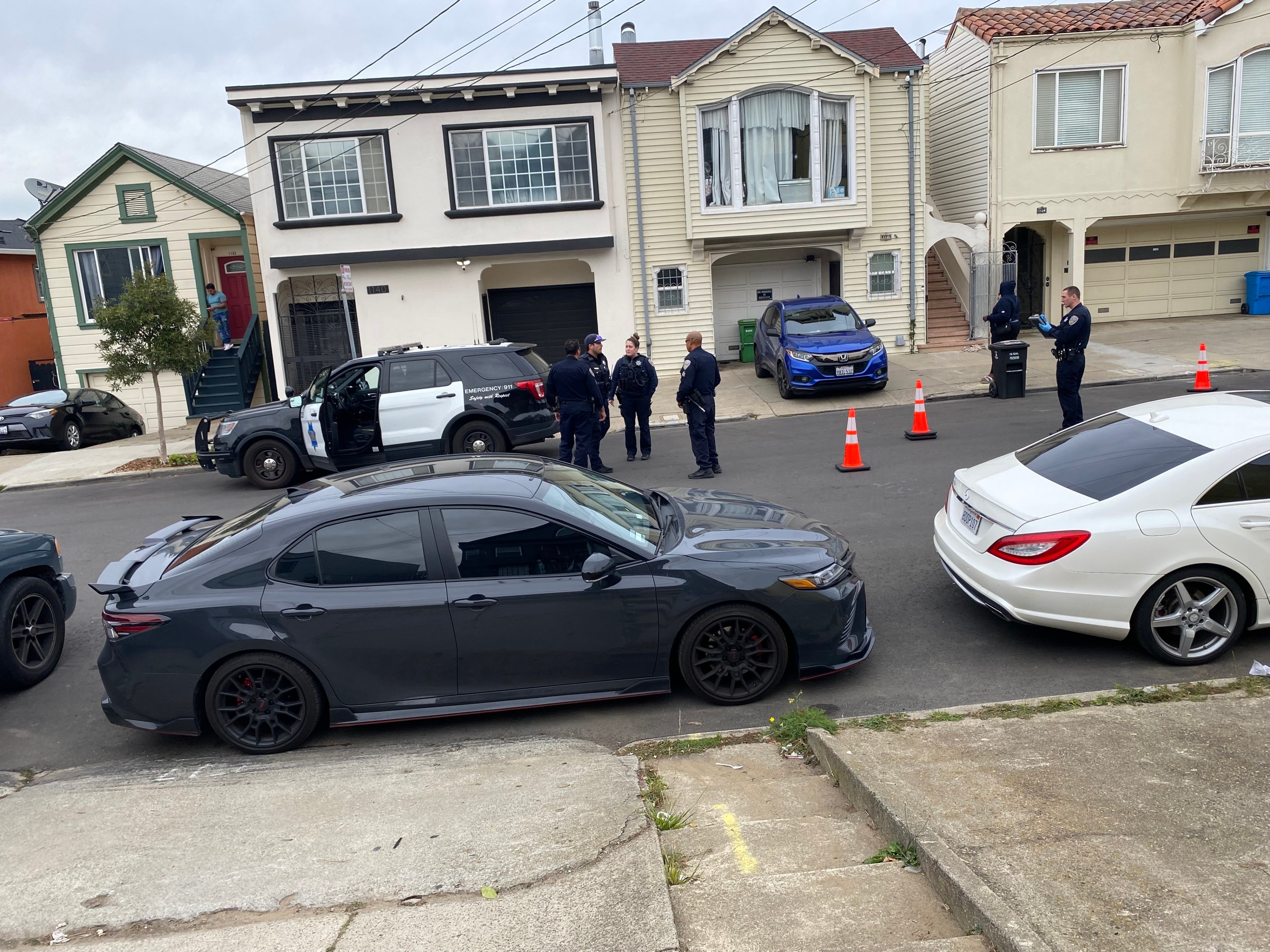 A group of police officers standing near their patrol car on a residential street. There are several cars and orange cones nearby, with houses in the background.