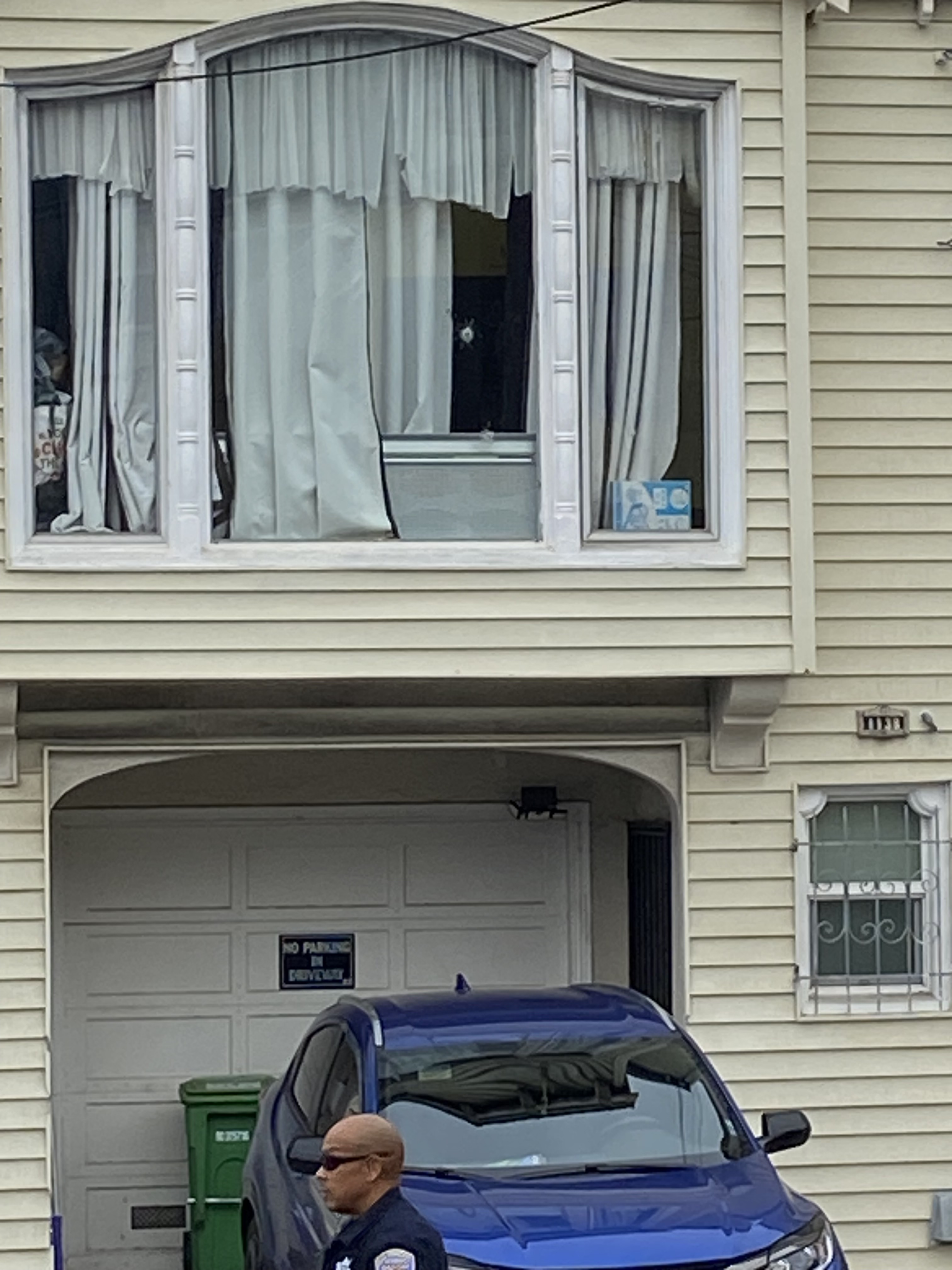 A man in uniform is standing in front of a blue car parked in a driveway with a &quot;No Parking&quot; sign. Above, a window with curtains partially open is visible.