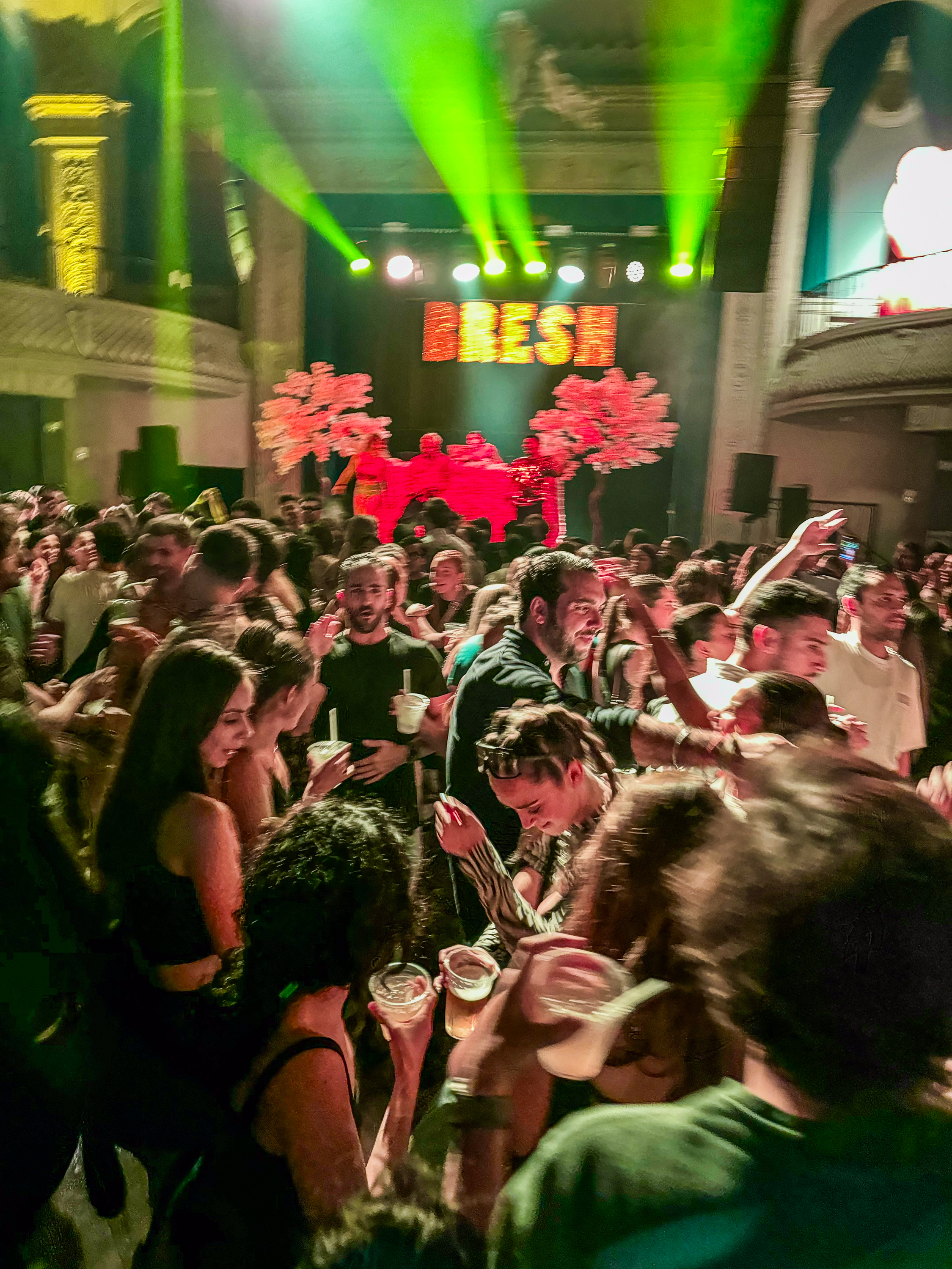 A lively nightclub scene shows a crowded dance floor with people holding drinks, colorful stage lights, and a &quot;FRESH&quot; sign in the background.