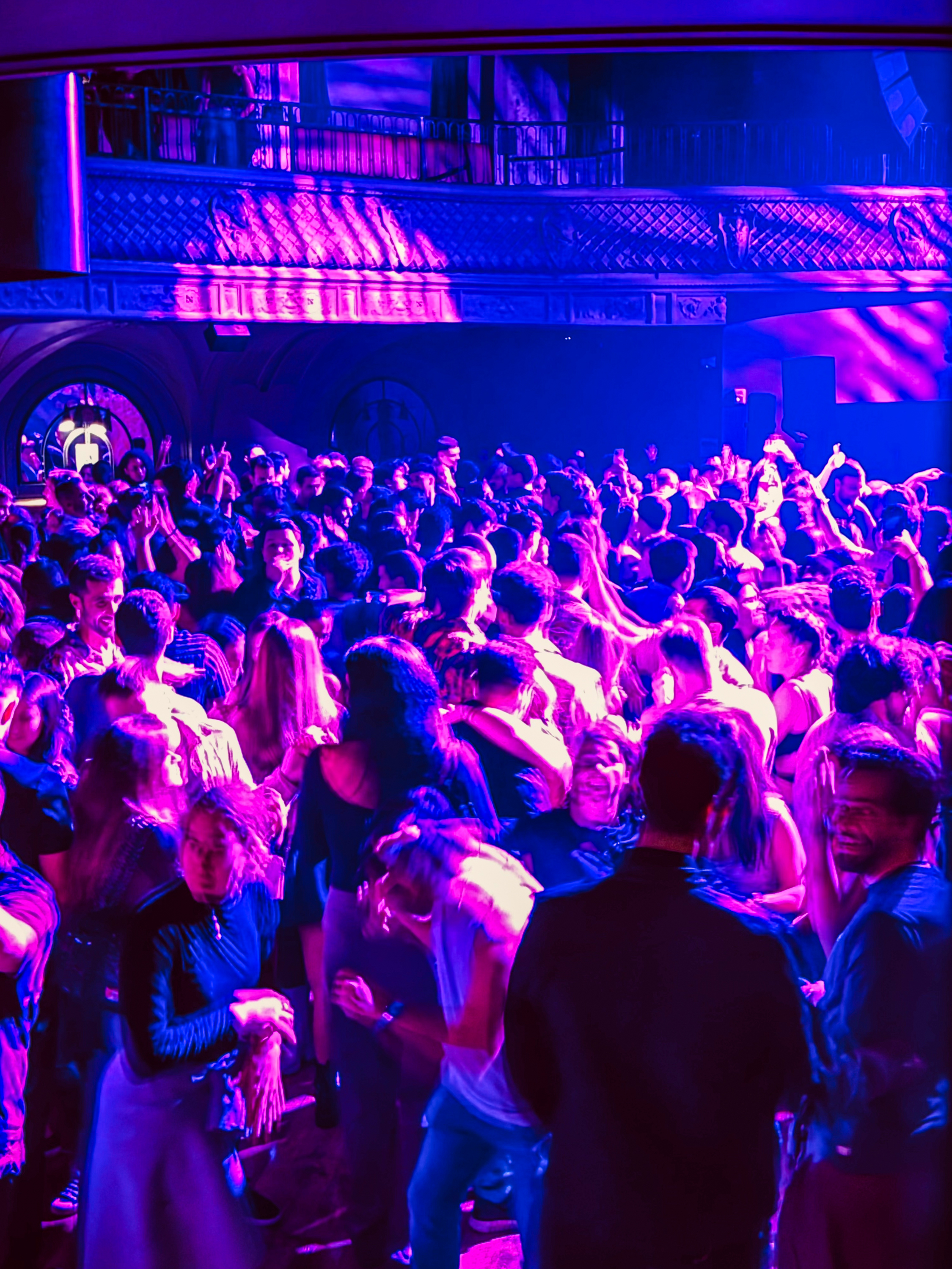 A large crowd of people is dancing in a vibrant nightclub, illuminated by bright purple and pink lighting. The scene is lively and energetic.