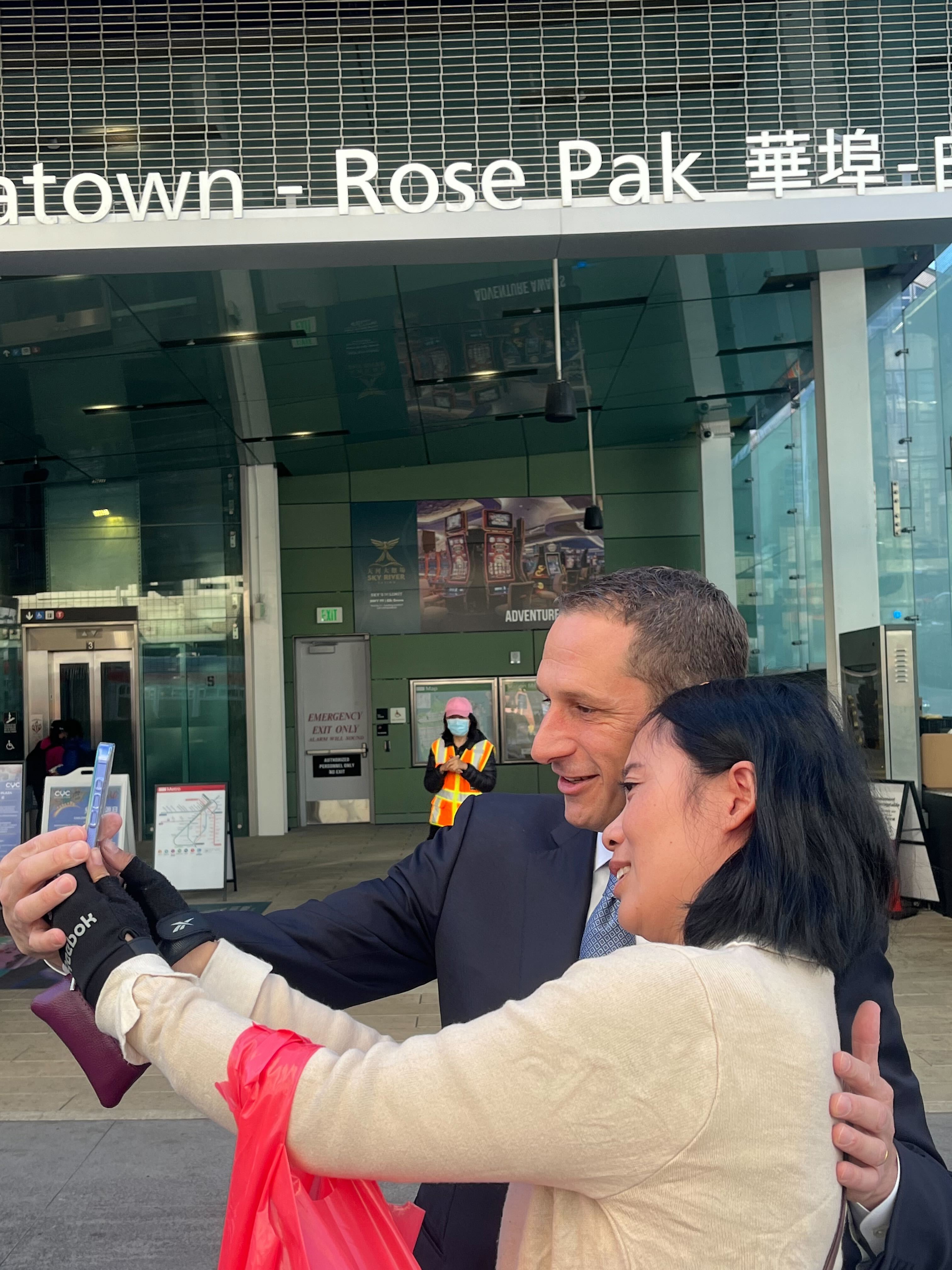 A man in a suit and a woman are taking a selfie together outside a building labeled &quot;Chinatown - Rose Pak&quot;. A person in a safety vest is in the background.