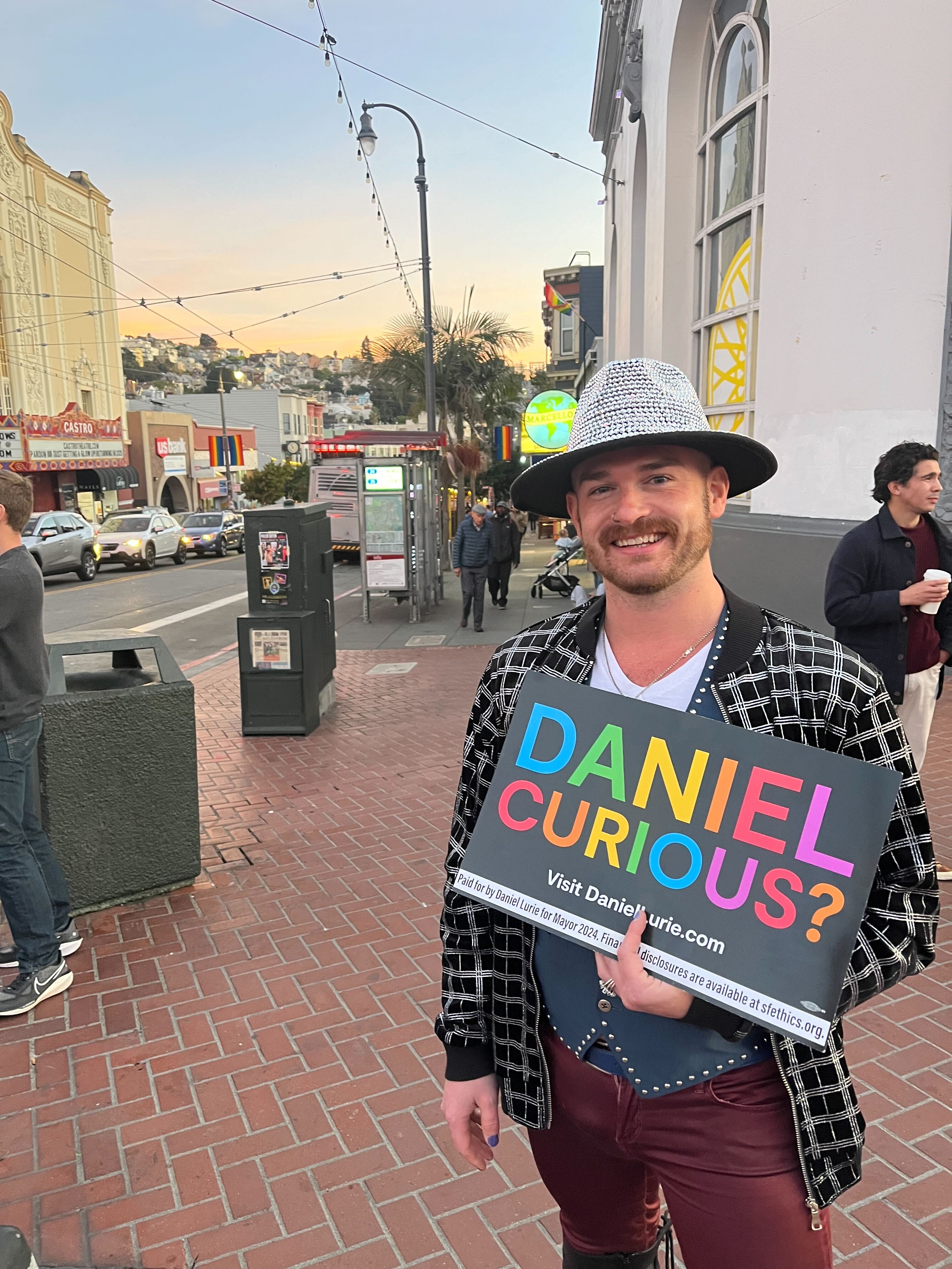 A person in a hat holds a colorful sign reading &quot;Daniel, curious?&quot; on a bustling city street with a theater, shops, and people in the background.