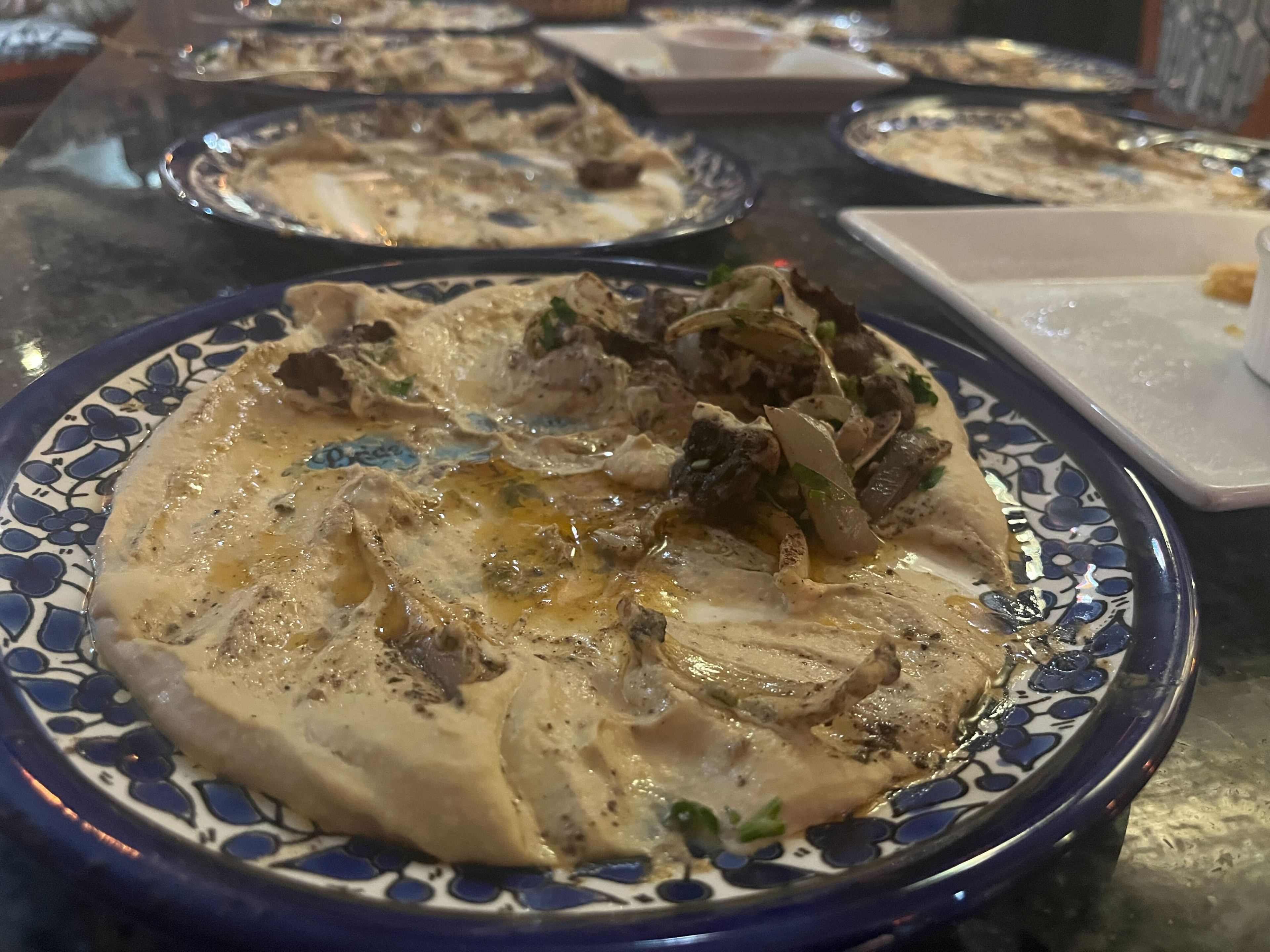 The image shows a decorative blue plate with creamy hummus topped with herbs and spices. Other similar plates are visible in the background on a dark table.