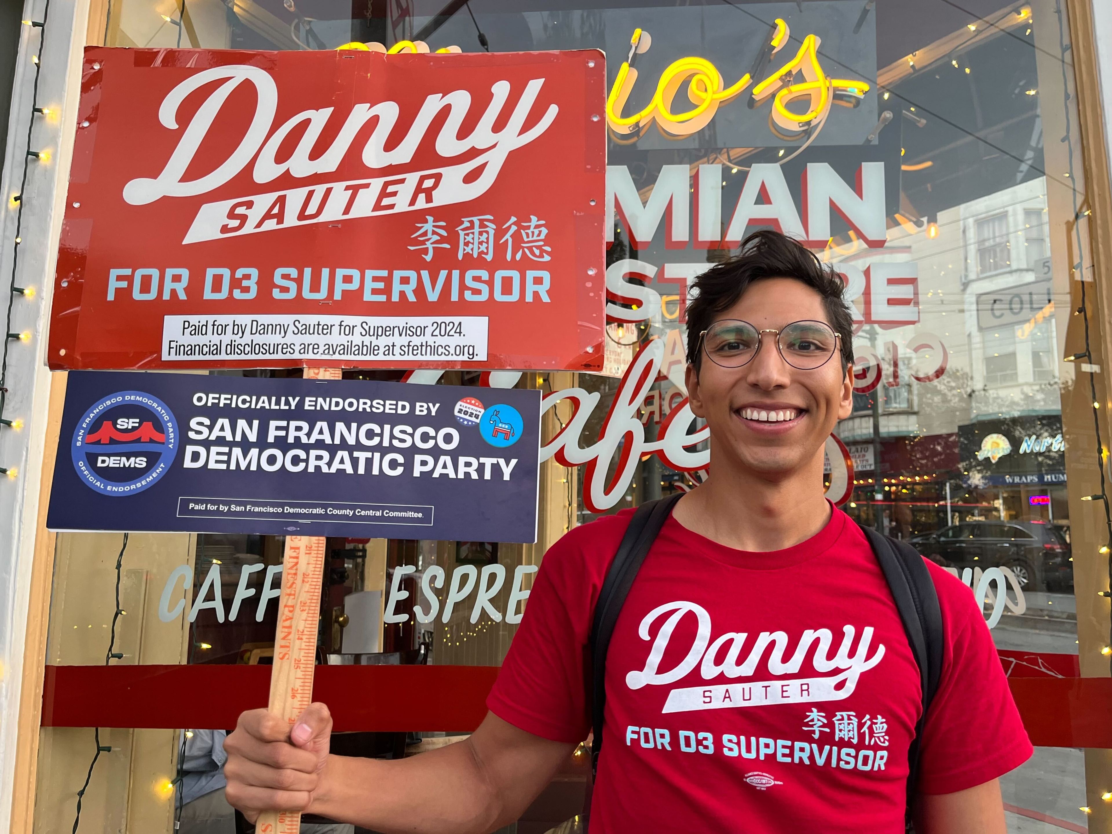 A smiling person in a red shirt holds campaign signs supporting Danny Sauter for D3 Supervisor, endorsed by the San Francisco Democratic Party, outside a café.