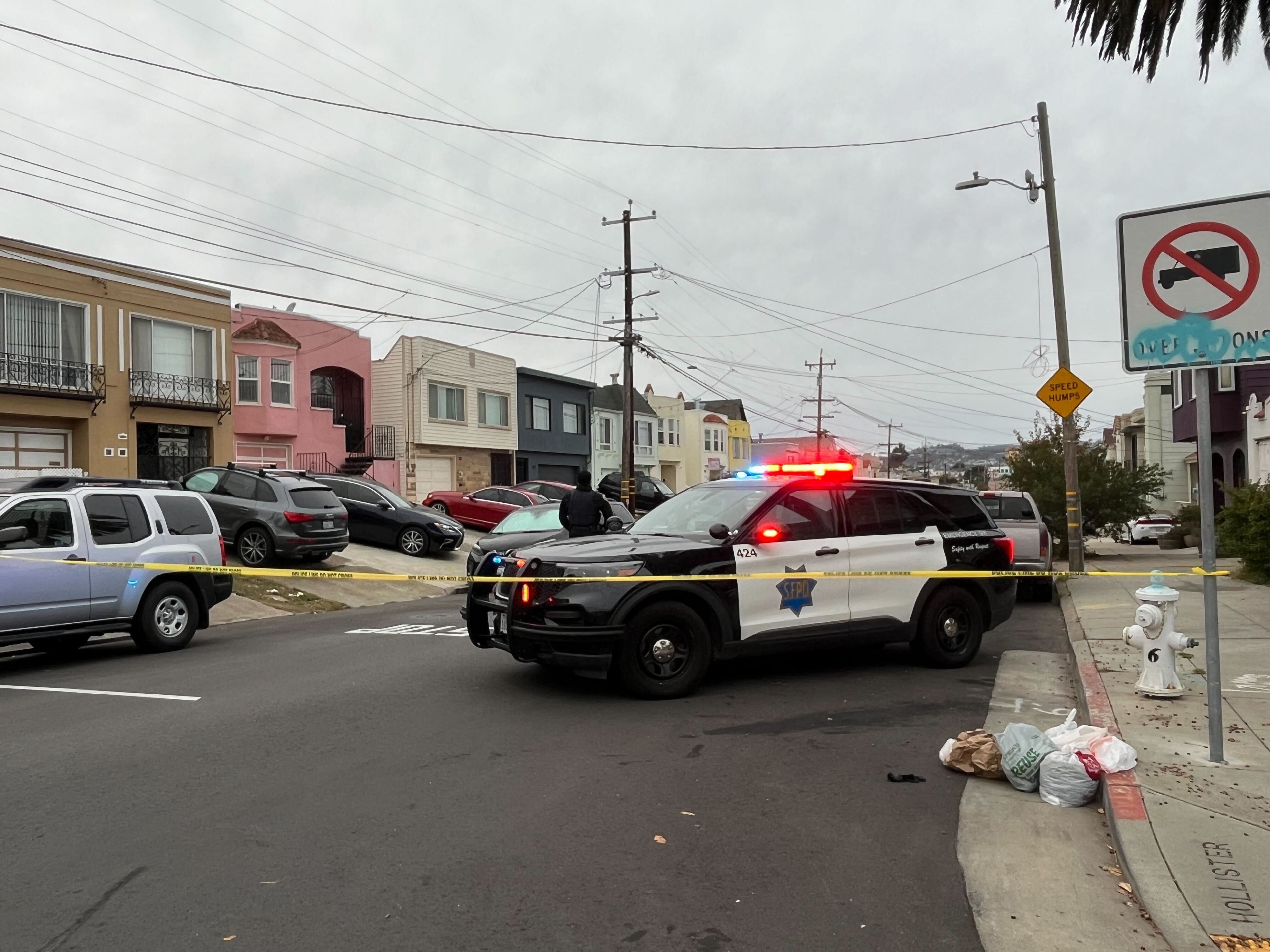 A police SUV with flashing lights blocks a street, alongside yellow caution tape. Nearby are parked cars, residential houses, and a few bags on the sidewalk.