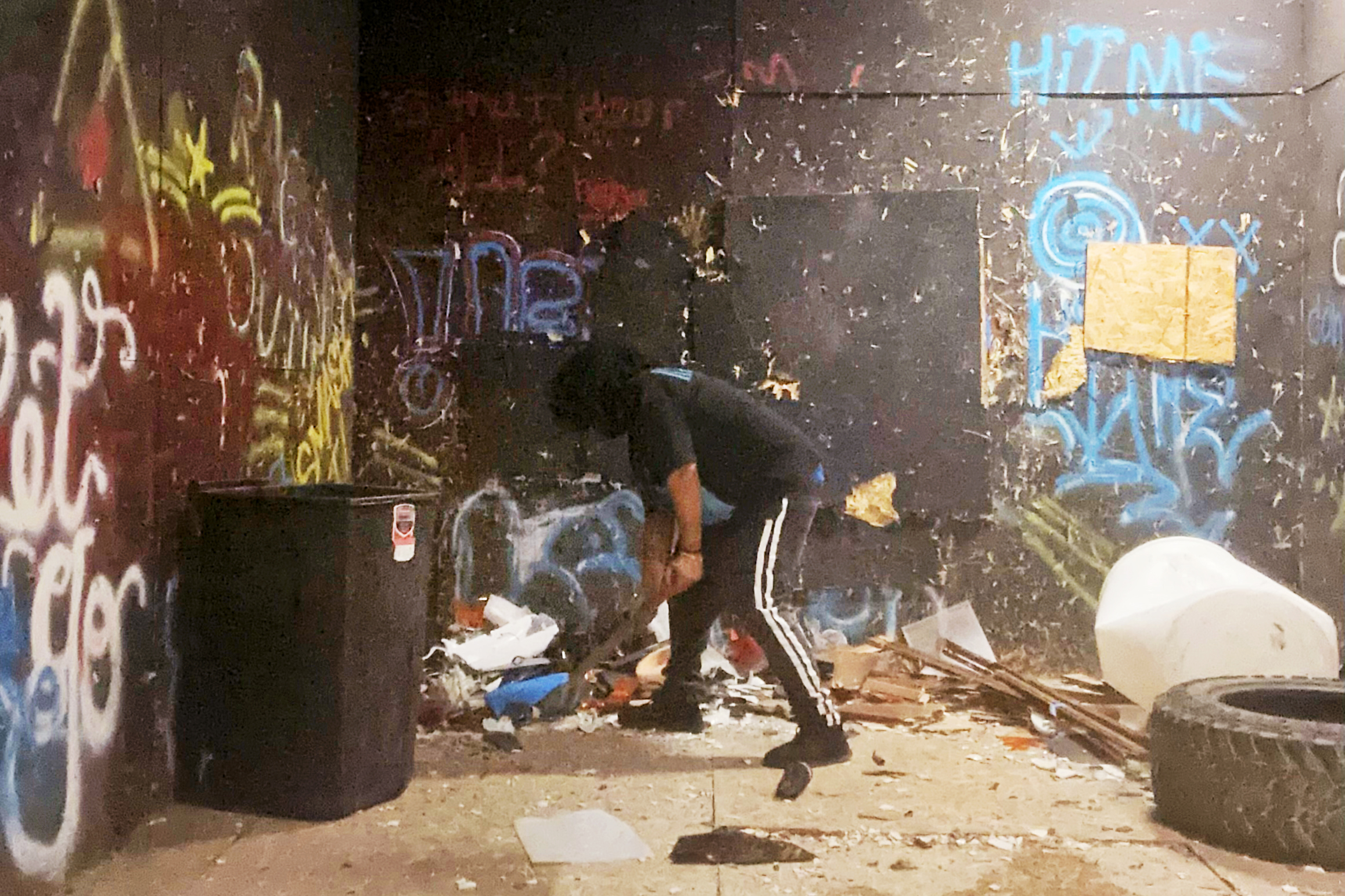 A person is smashing debris in a dimly lit room with graffiti-covered walls. The floor is littered with trash, and a large tire is on the right.