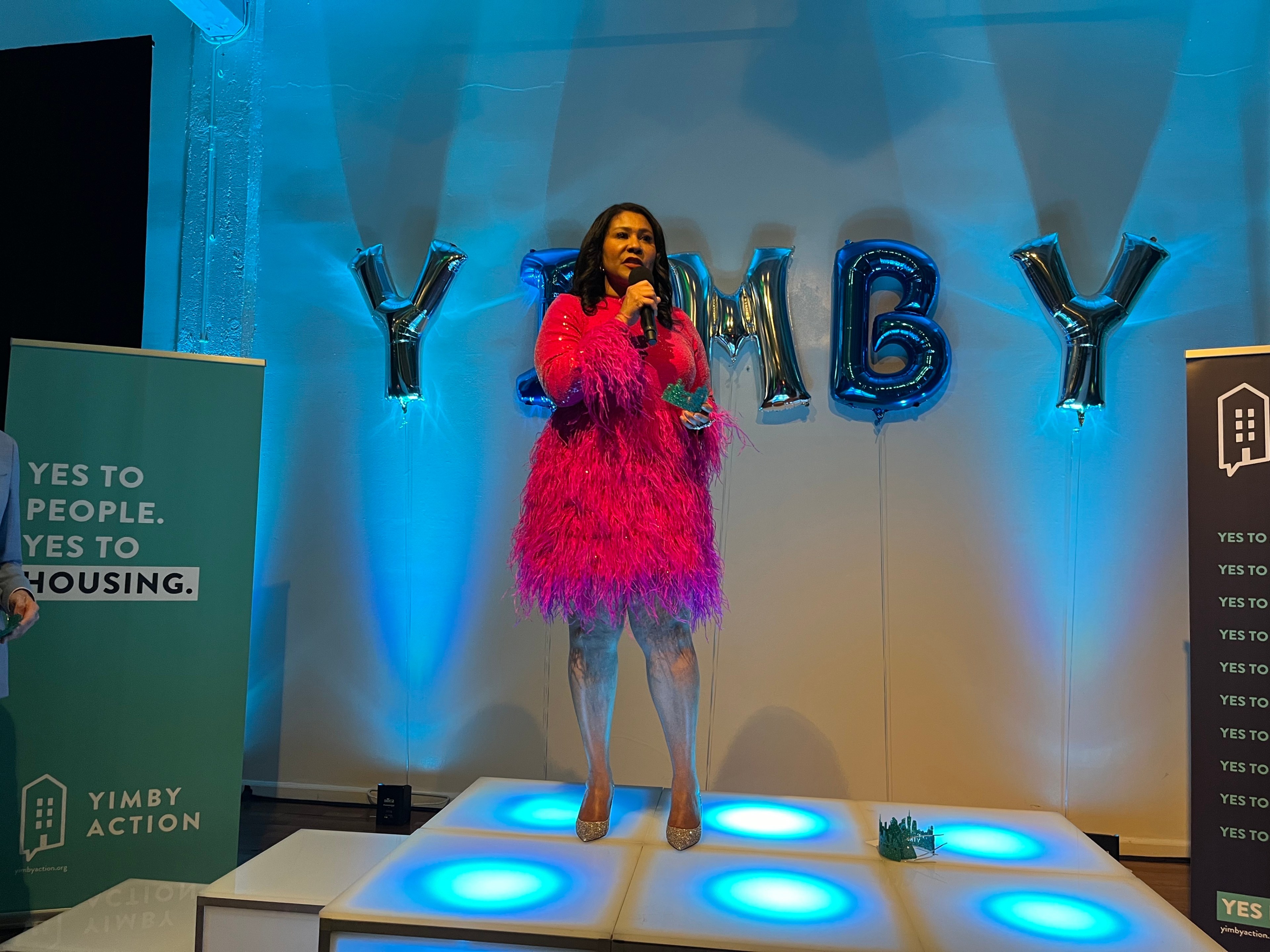 Mayor Breed in a puffy pink dress stands on a dance floor with blue lights and balloons spelling YIMBY in the background.