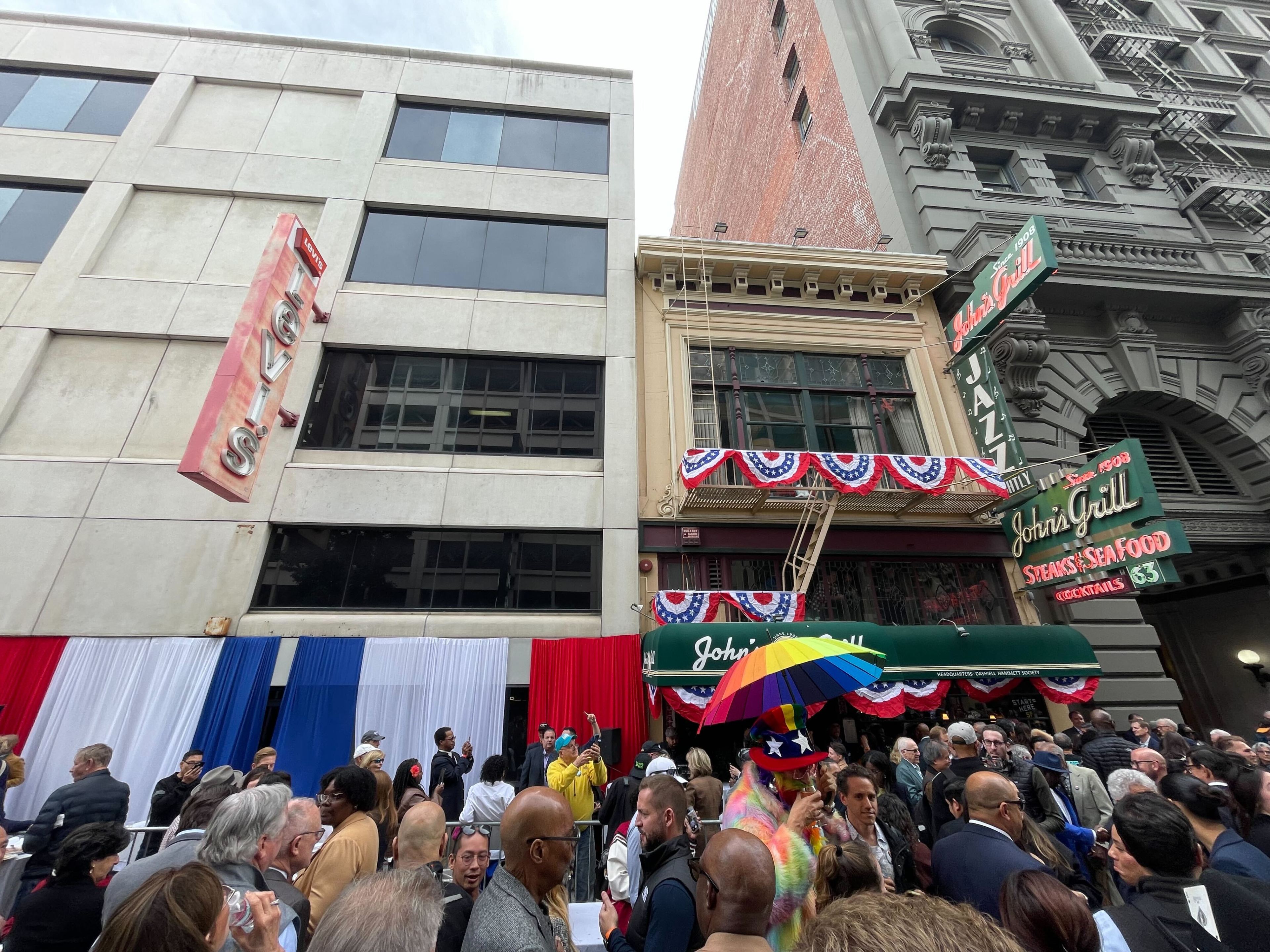 A lively crowd gathers outside buildings with ornate architecture, decorated with patriotic bunting. A person holds a colorful umbrella in the foreground.