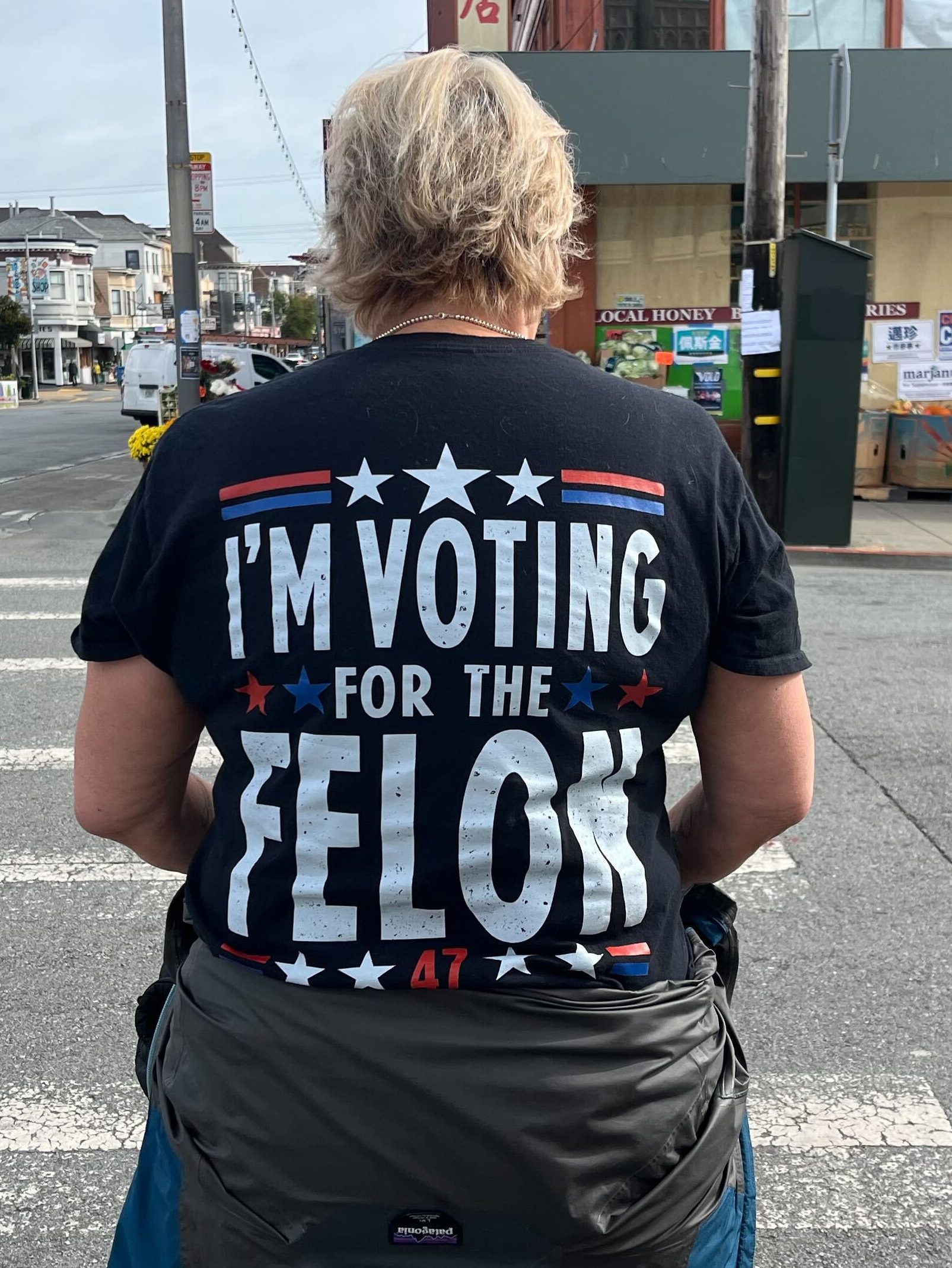 A person is standing at a crosswalk wearing a black T-shirt that says &quot;I'M VOTING FOR THE FELON&quot; in white letters. The background shows a street scene.