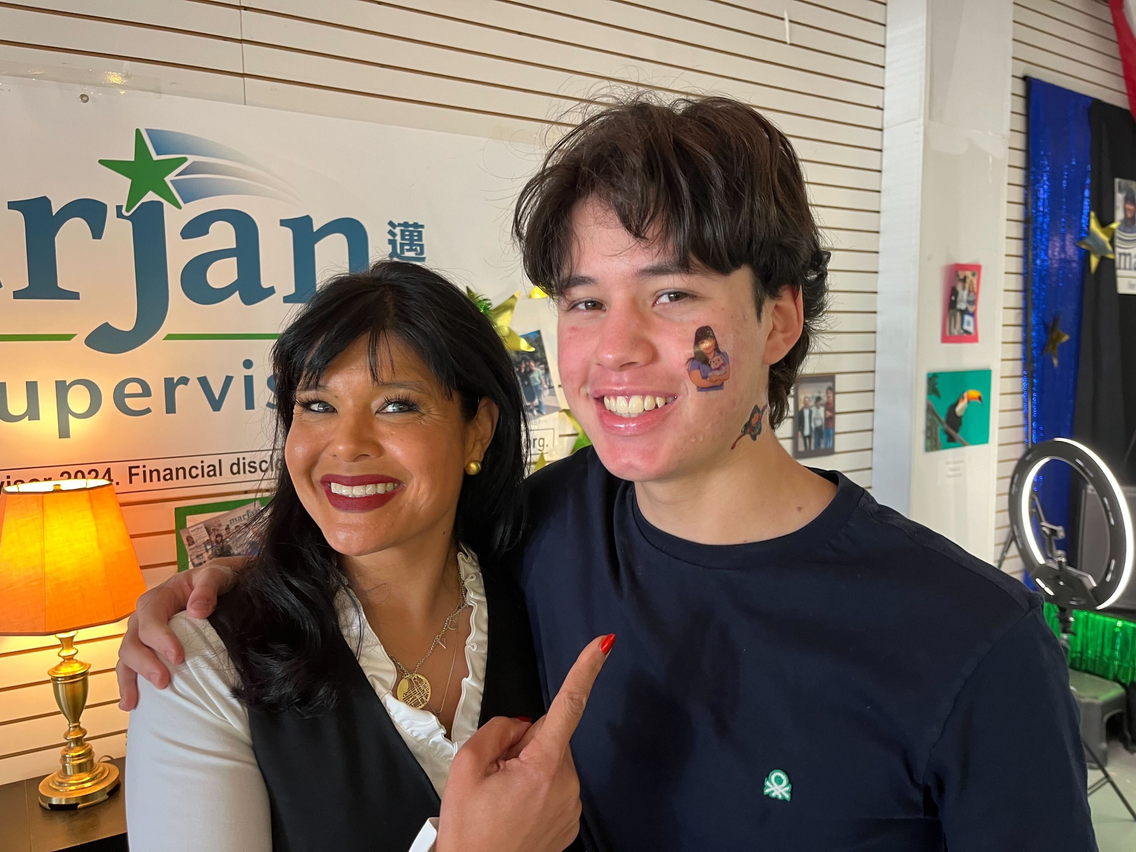 A woman with dark hair smiles and points at a young man with a face tattoo. They stand together, inside a room with a lit lamp and various photos on the wall.