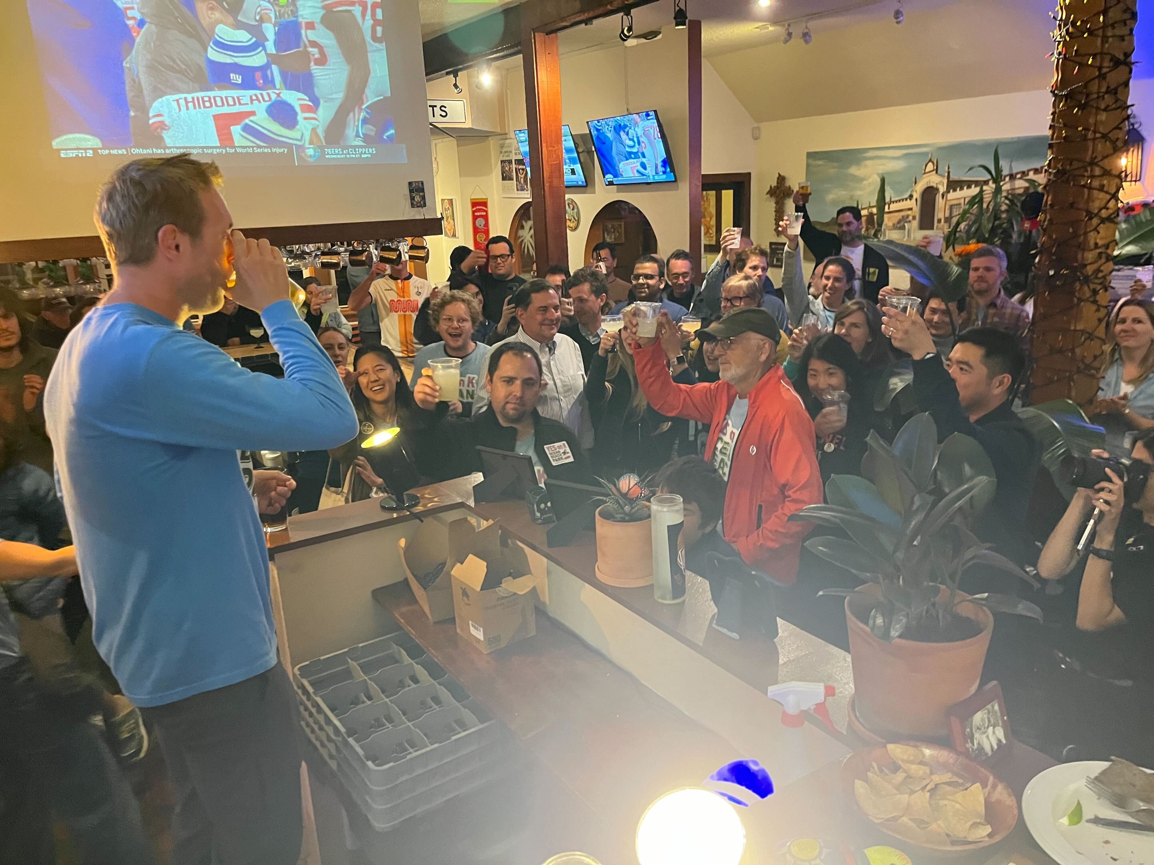 A man in a blue shirt drinks from a glass as a cheering crowd raises their glasses. They are gathered in a bar with a sports game projected on the wall.