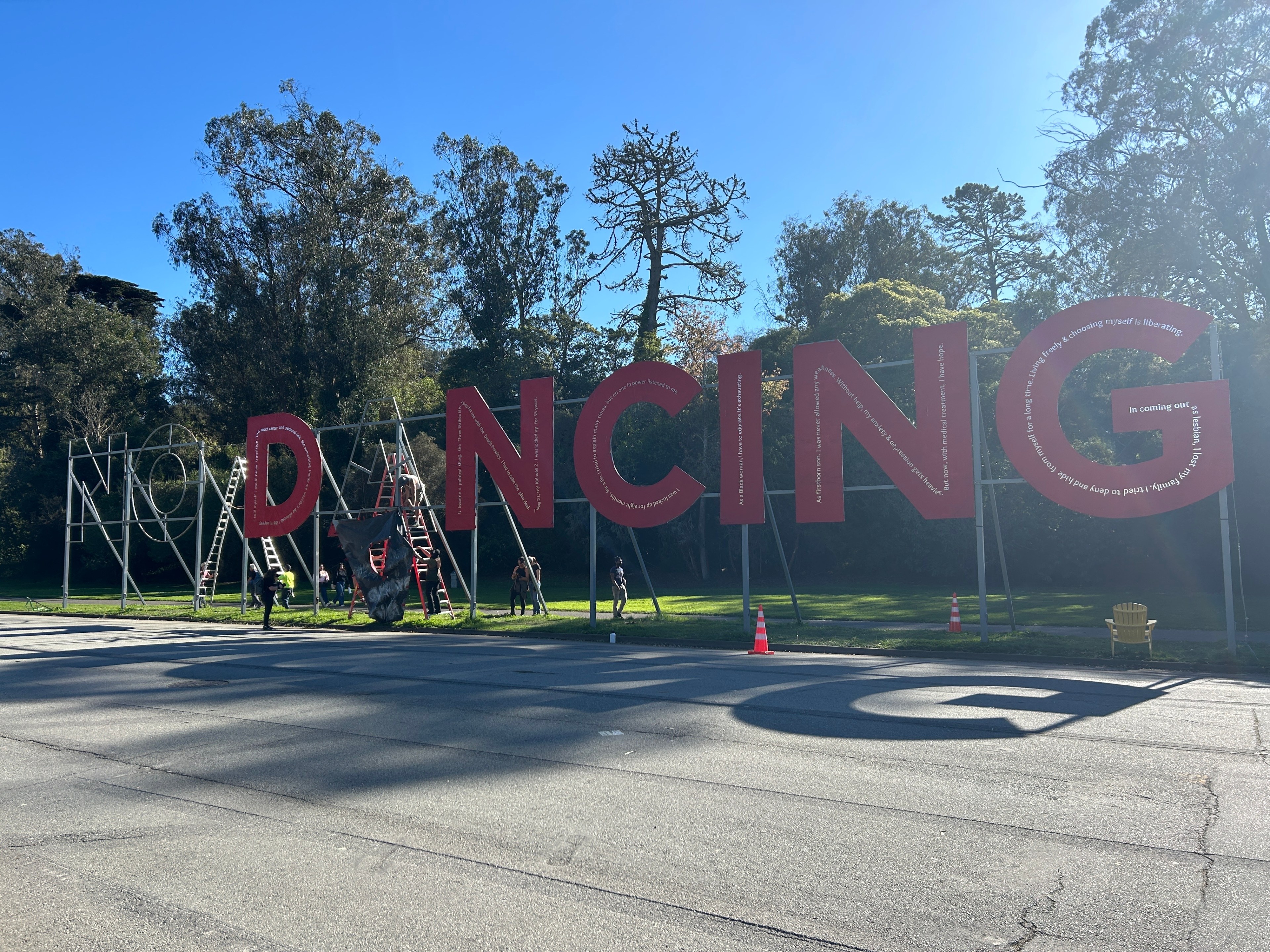 The image shows a large sign spelling &quot;NO DANCING&quot; in red letters, with people working on it using ladders. It's set against a backdrop of tall trees and a blue sky.