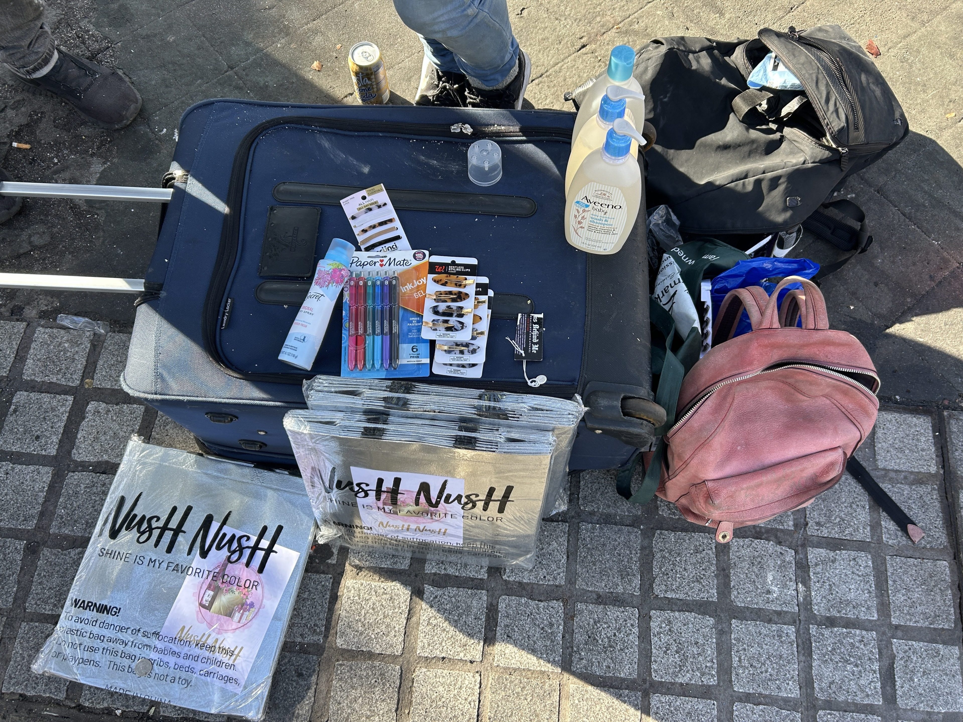 The image shows a suitcase with stationery, hair clips, and a tube on top. Nearby, there's a black backpack, a pink bag, and three bottles of lotion.