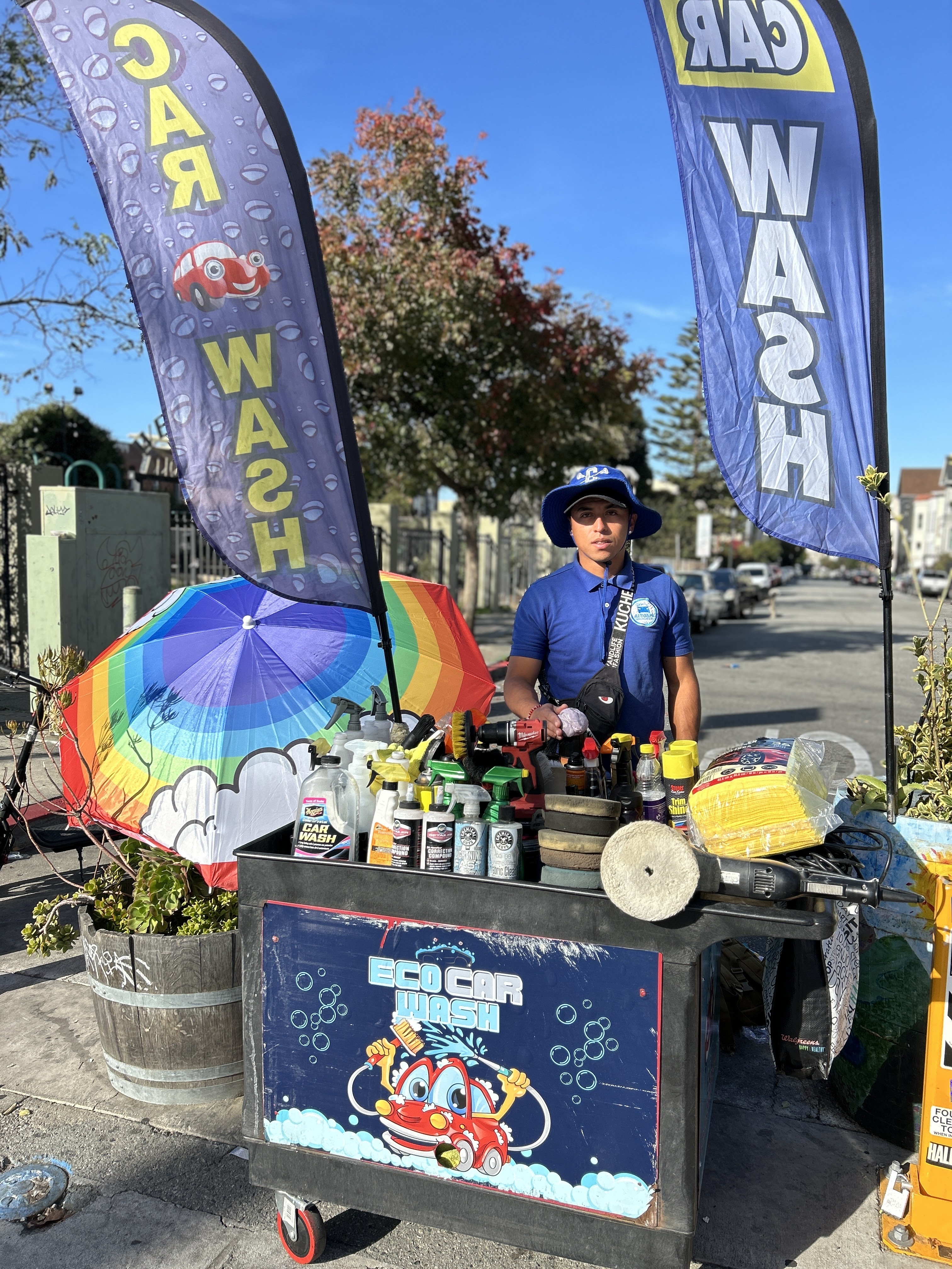 A person stands behind an &quot;Eco Car Wash&quot; cart filled with cleaning supplies. Two large banners read &quot;CAR WASH&quot; beside a colorful umbrella on a sunny street.