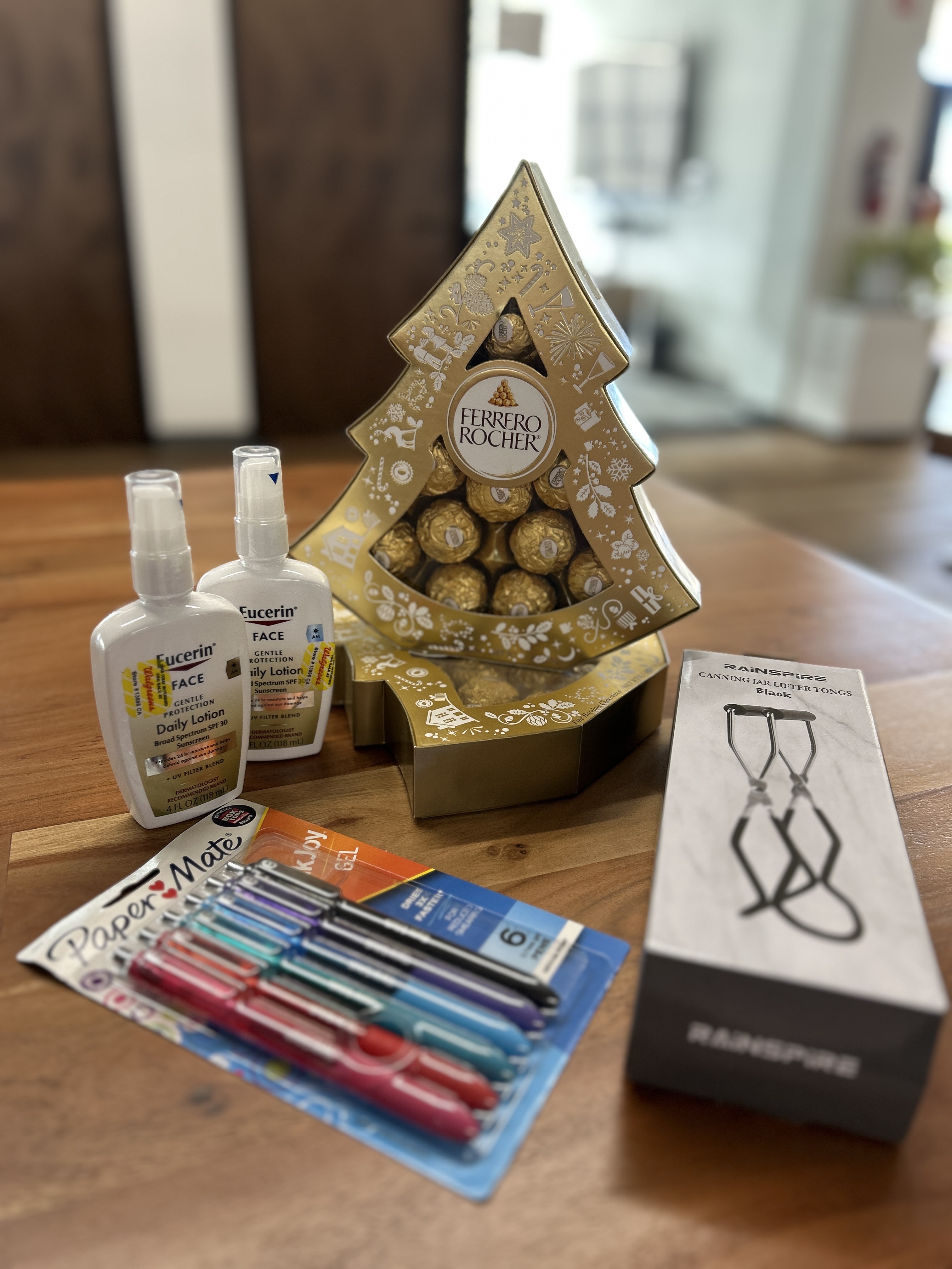 The image shows a gold tree-shaped Ferrero Rocher box, two Eucerin lotions, a pack of colorful Paper Mate pens, and a box of black canning tongs on a wooden table.