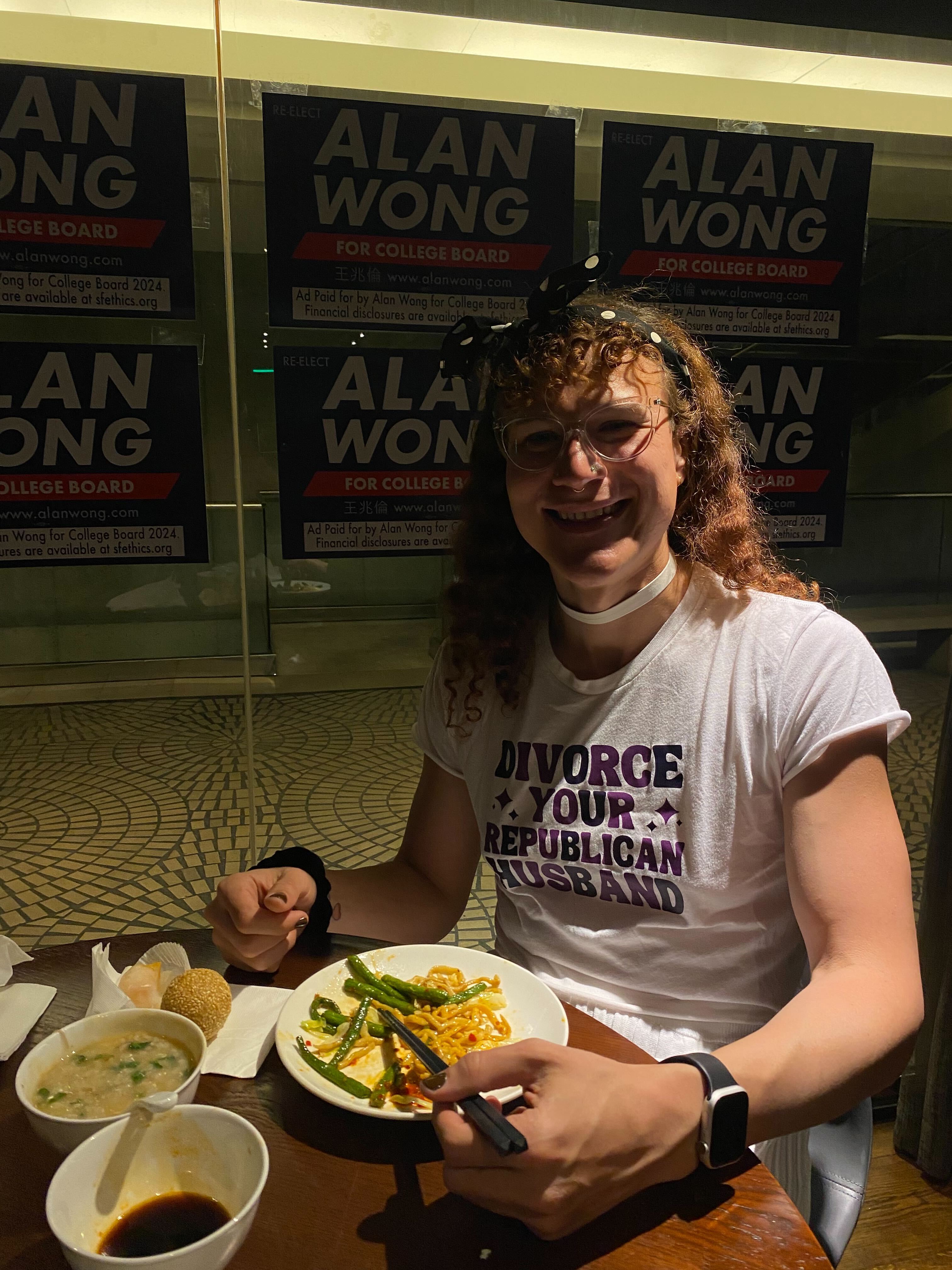 A person wearing a T-shirt with a slogan is seated at a table with various dishes, including noodles and soup, in front of campaign posters for Alan Wong.