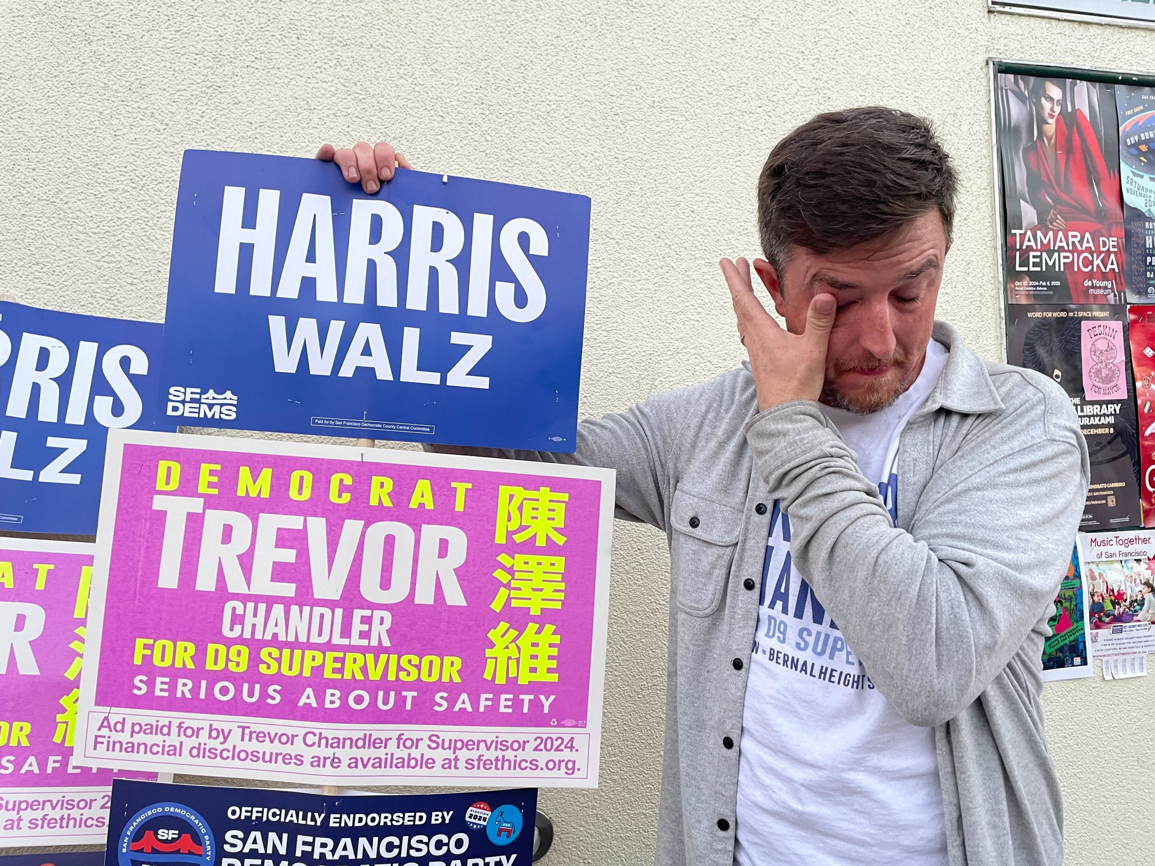 A man stands by a wall holding campaign signs for Harris Walz and Trevor Chandler, while wiping his eye. There's a poster behind him on the wall.