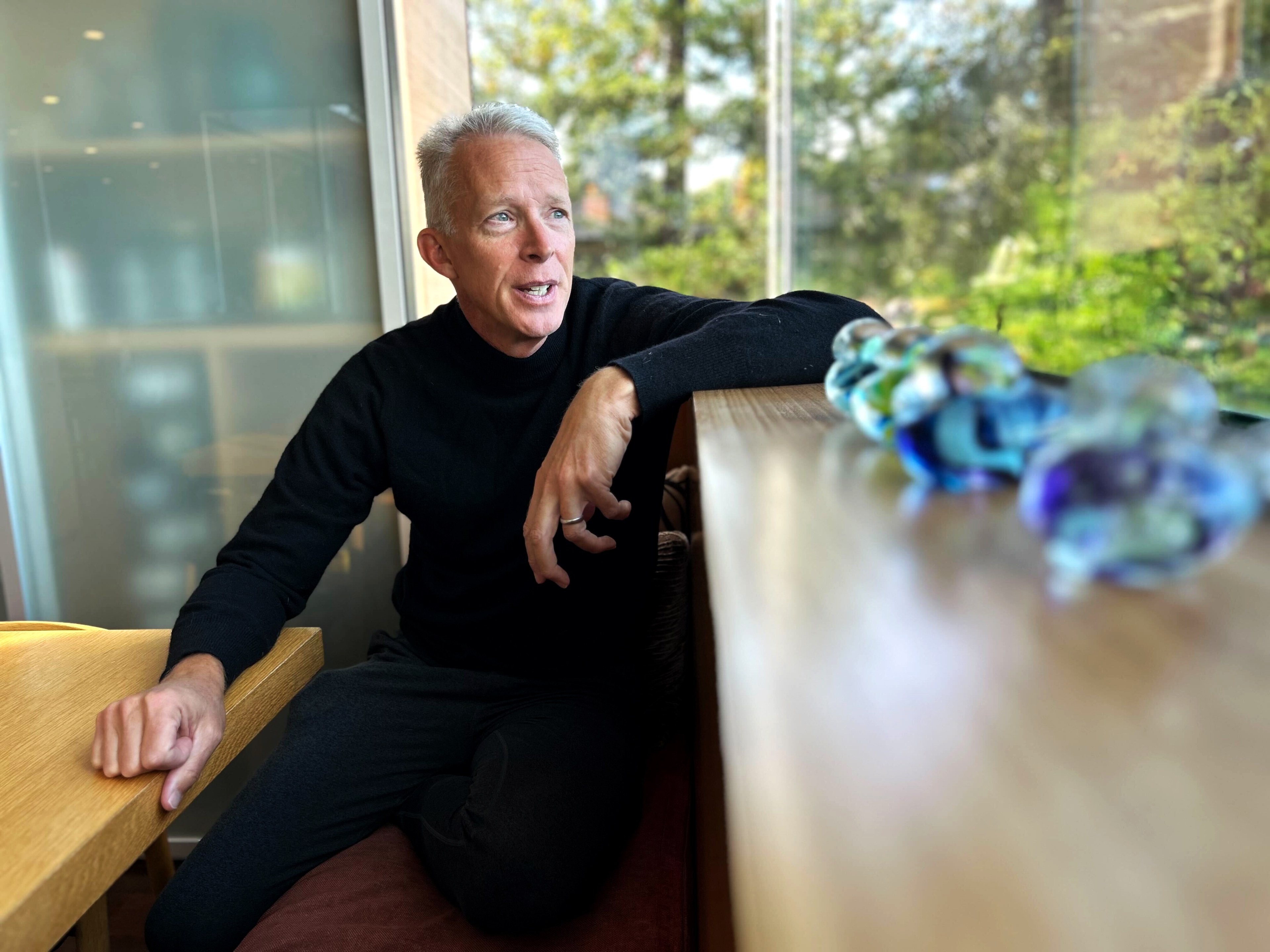 A man in a black sweater sits by a table, gazing out a large window with trees outside. Colorful glass pebbles are blurred in the foreground.