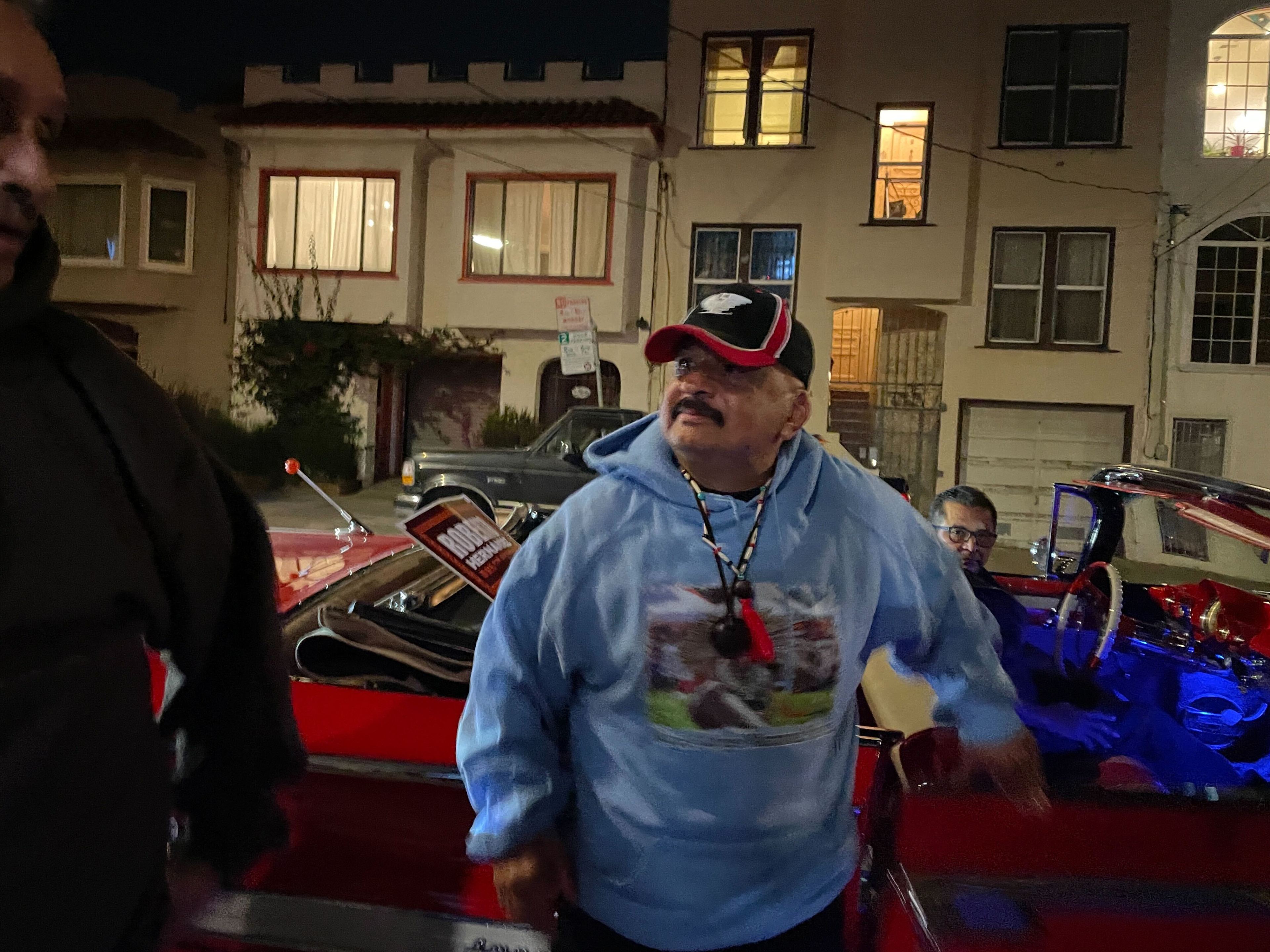 A man in a light blue hoodie and cap stands beside a red car at night, with another person seated inside. Residential buildings are in the background.