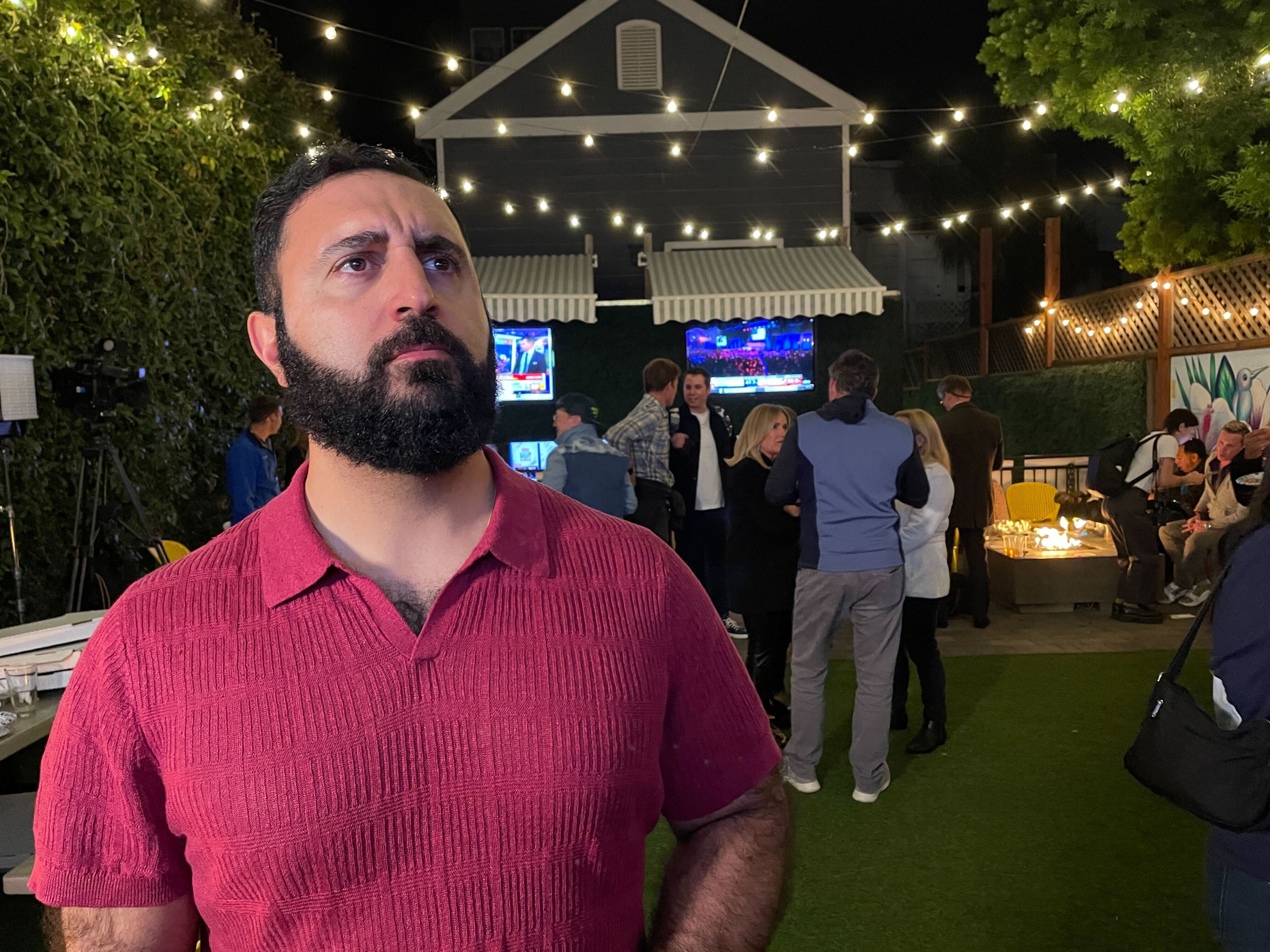 A bearded man in a red shirt looks contemplative at an outdoor gathering with string lights, people chatting, and two TVs in the background showing sports.