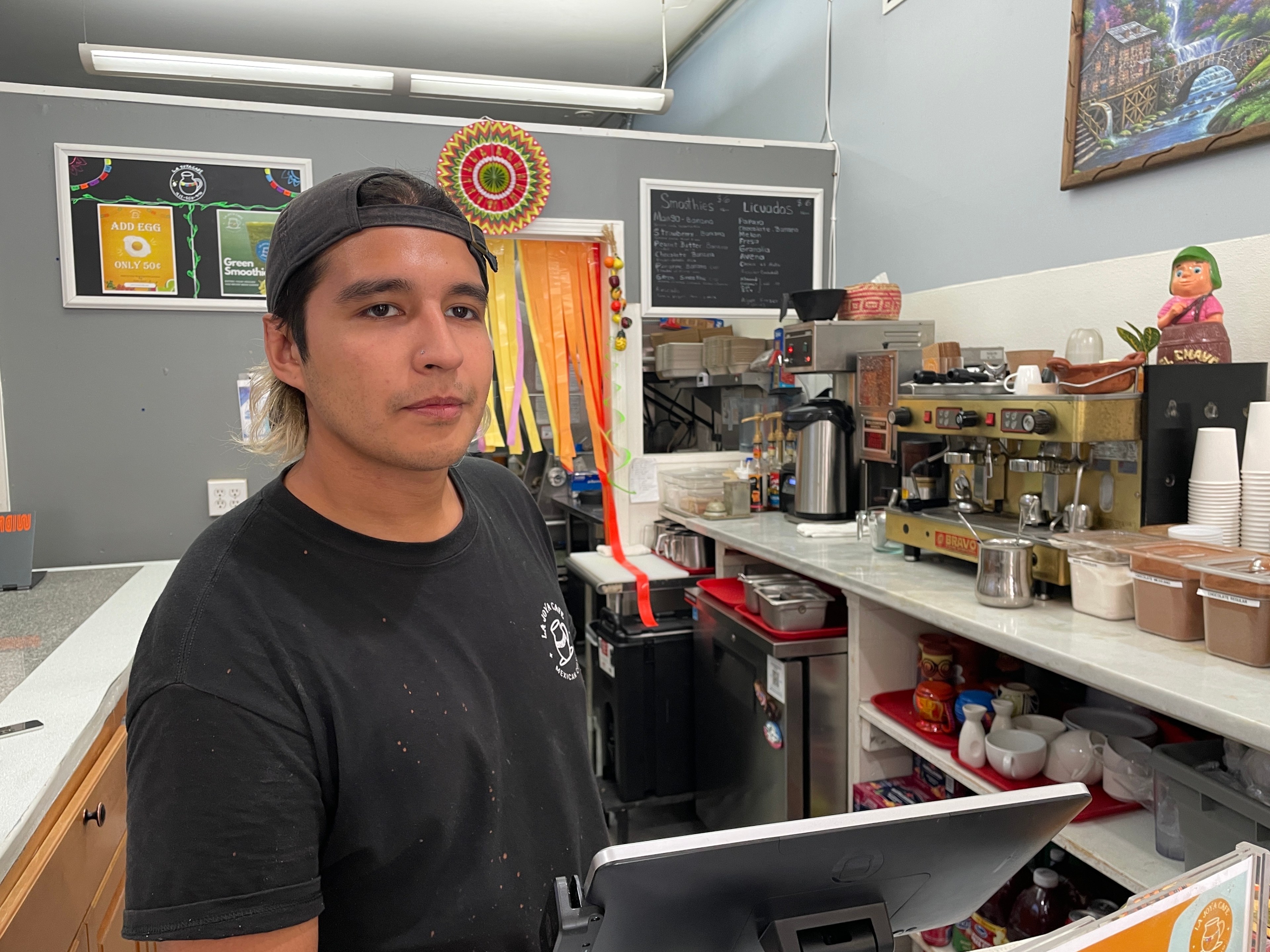 A person in a black shirt and cap stands behind a counter in a café. The counter has a coffee machine, cups, and storage containers. Behind them are colorful decorations.