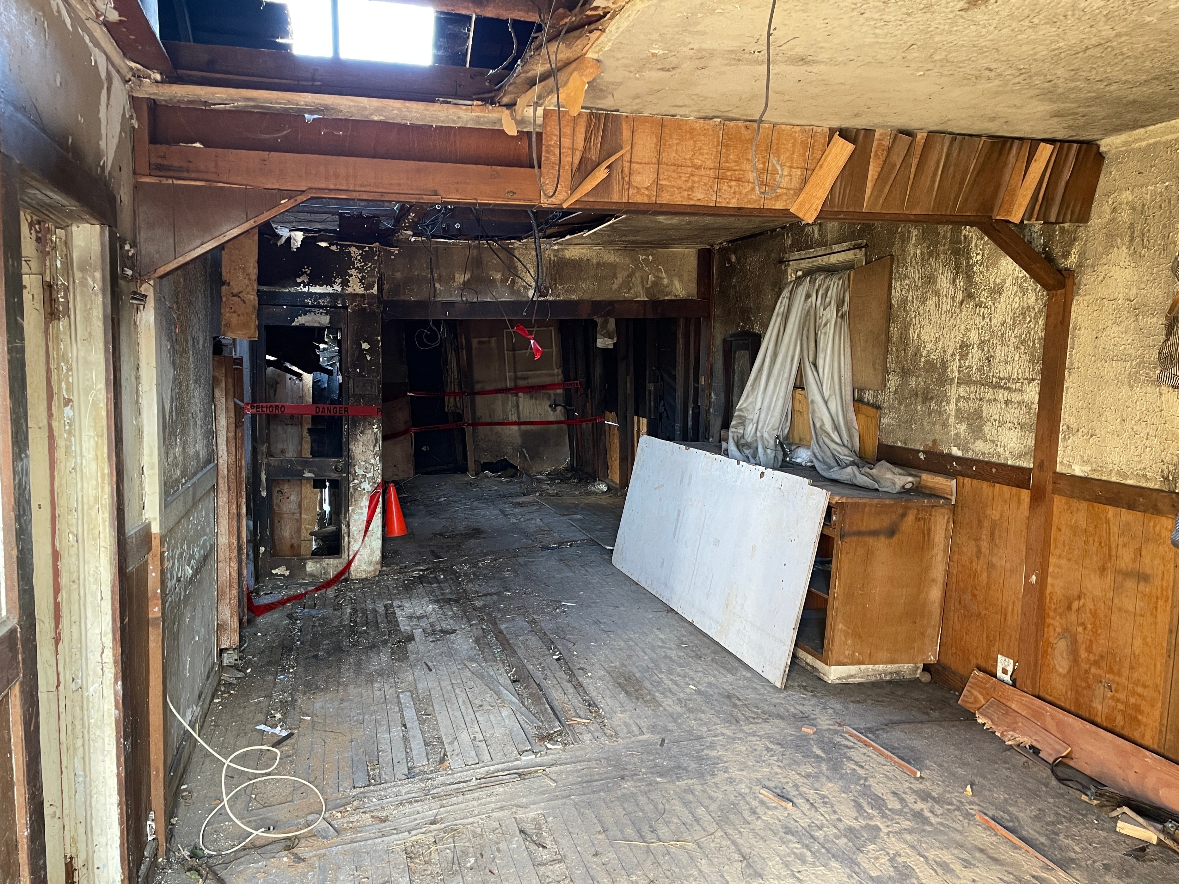 The image shows a damaged, abandoned room with exposed beams, debris on the wooden floor, red caution tape, hanging wires, and a partially collapsed ceiling.