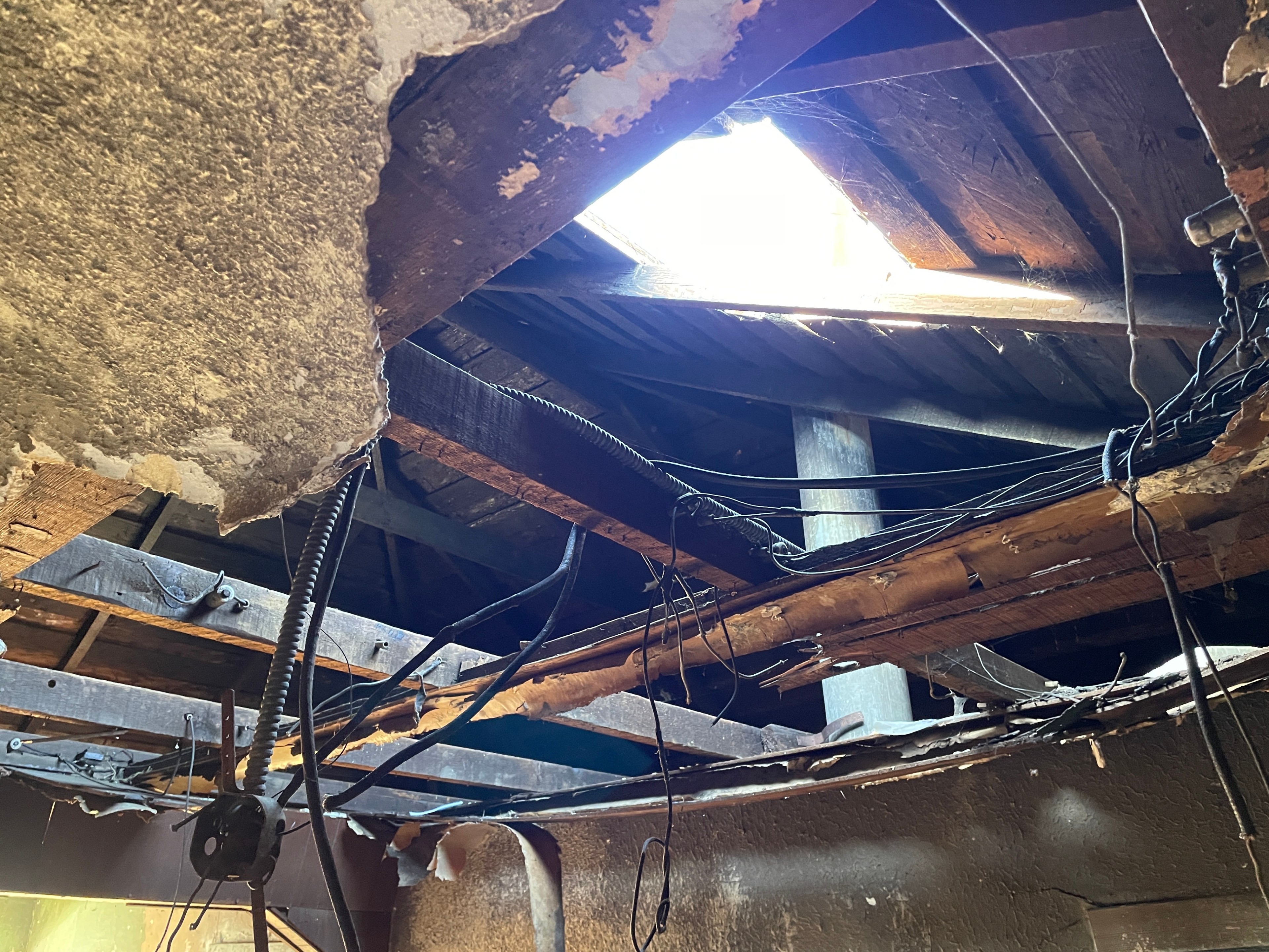 The image shows a damaged ceiling with exposed wooden beams and electrical wires. A hole lets sunlight in, illuminating the worn, crumbling structure.