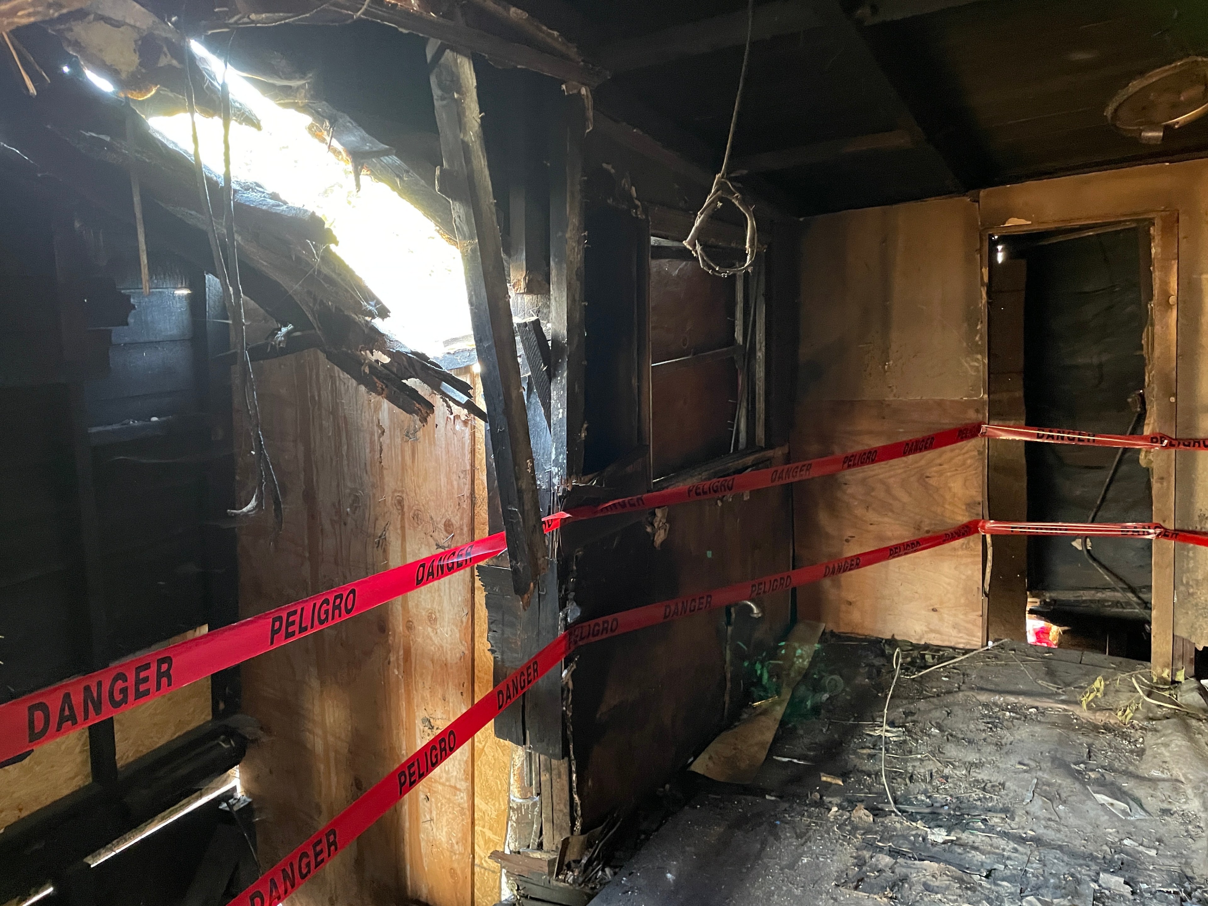 The image shows a charred, damaged room with blackened walls and a partially collapsed ceiling. Red &quot;Danger&quot; tape is strung around, cordoning off the area.