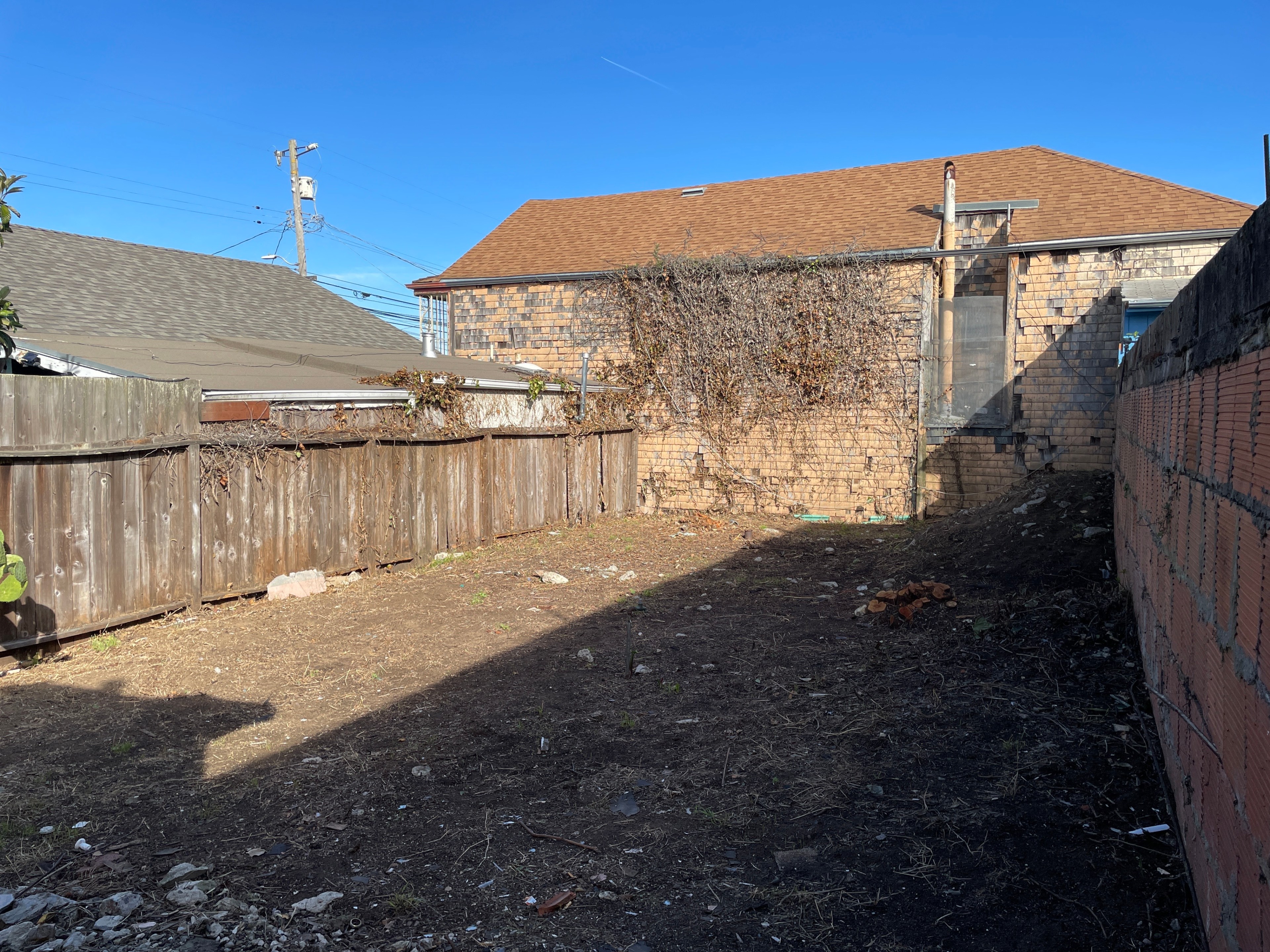 A small, barren dirt yard is bordered by a wooden fence and a brick wall with vines. An adjacent building features a brown shingle roof under a clear blue sky.