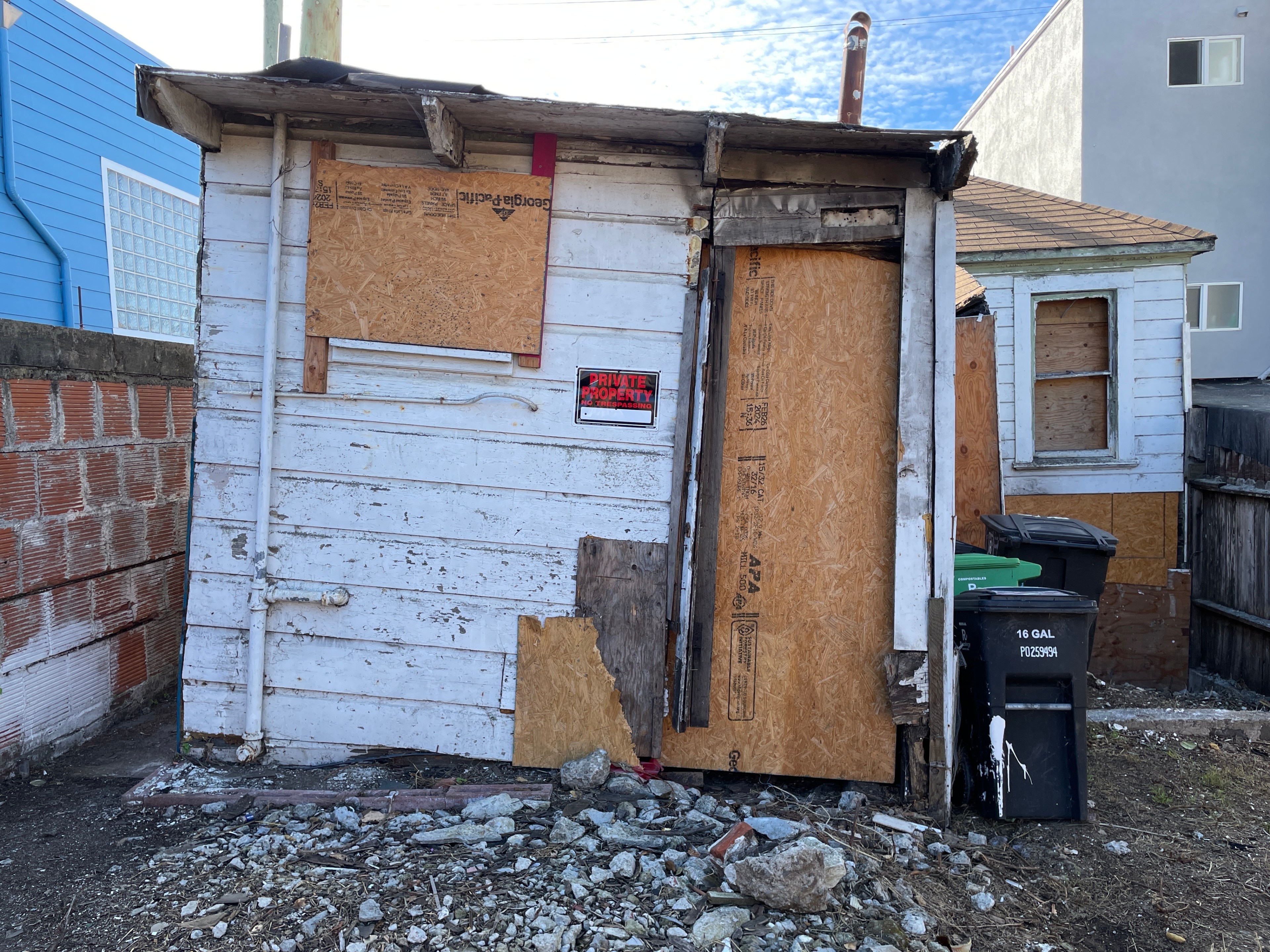 A small, weathered wooden shack with boarded-up windows and doors is surrounded by debris. A “Private Property” sign is displayed, and trash bins sit nearby.