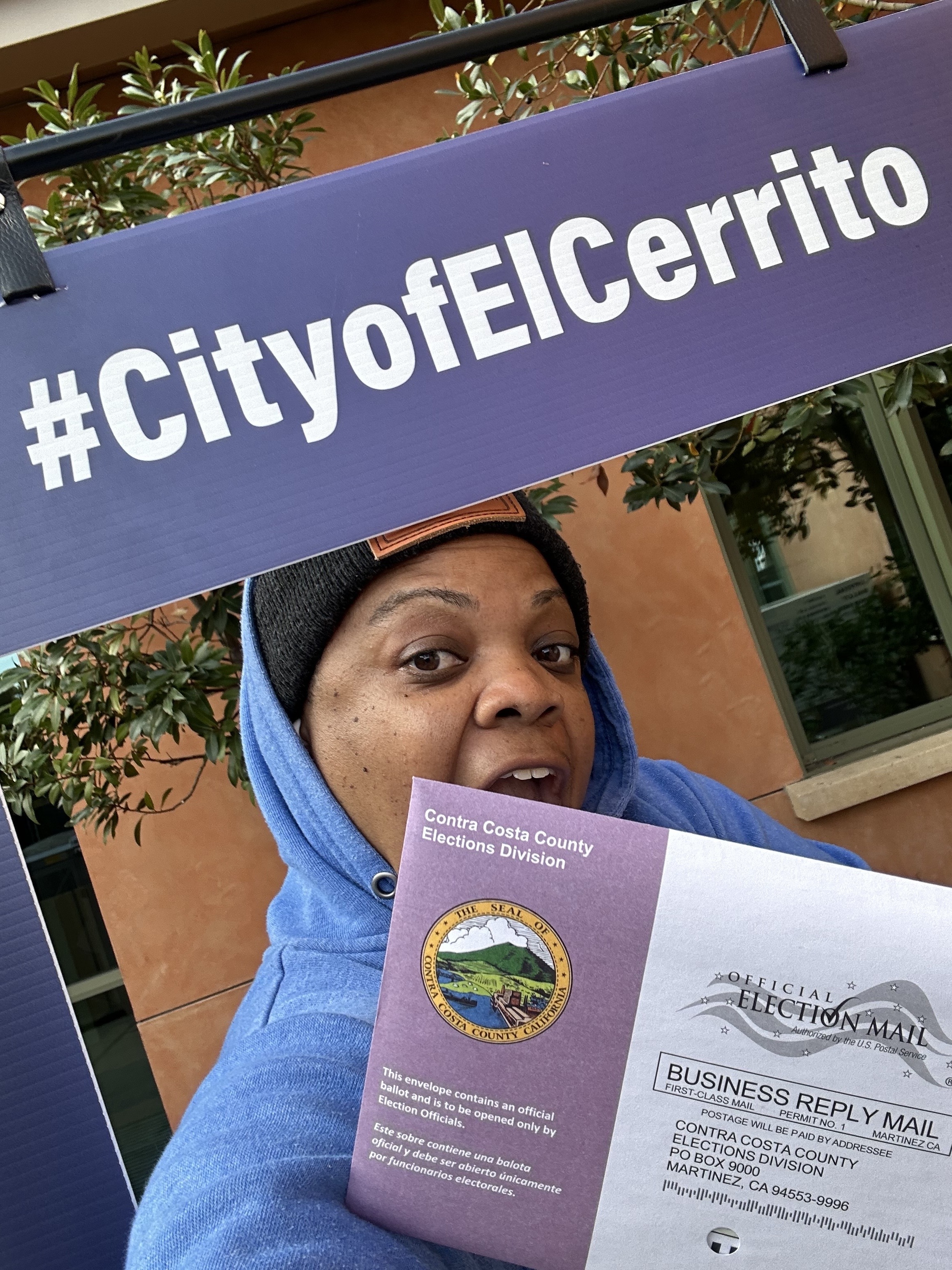 A person in a blue hoodie holds an election ballot envelope labeled &quot;Contra Costa County Elections Division&quot; under a #CityofElCerrito sign.