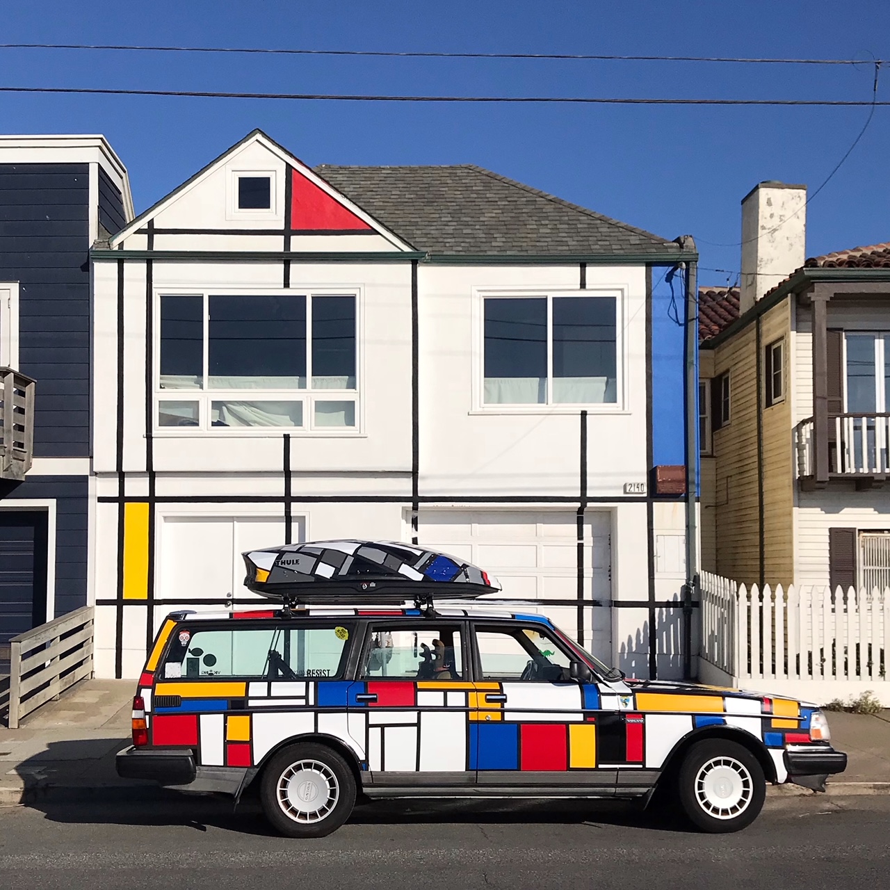 A car and a house are painted in a Mondrian style with geometric patterns using black lines and blocks of red, yellow, and blue on white surfaces.