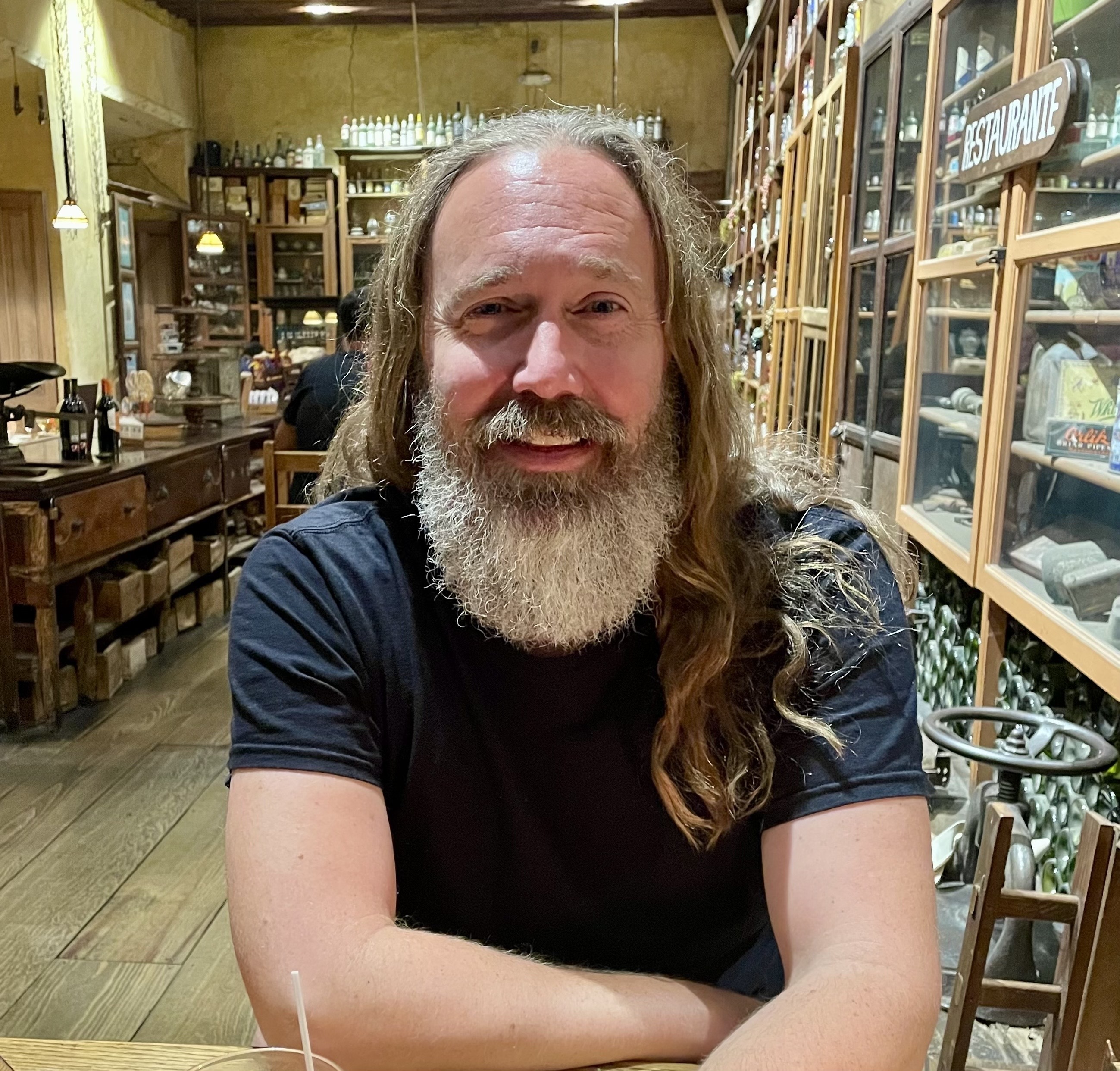 A man with long hair and a beard sits smiling in a rustic restaurant with wooden furnishings and shelves lined with various bottles.