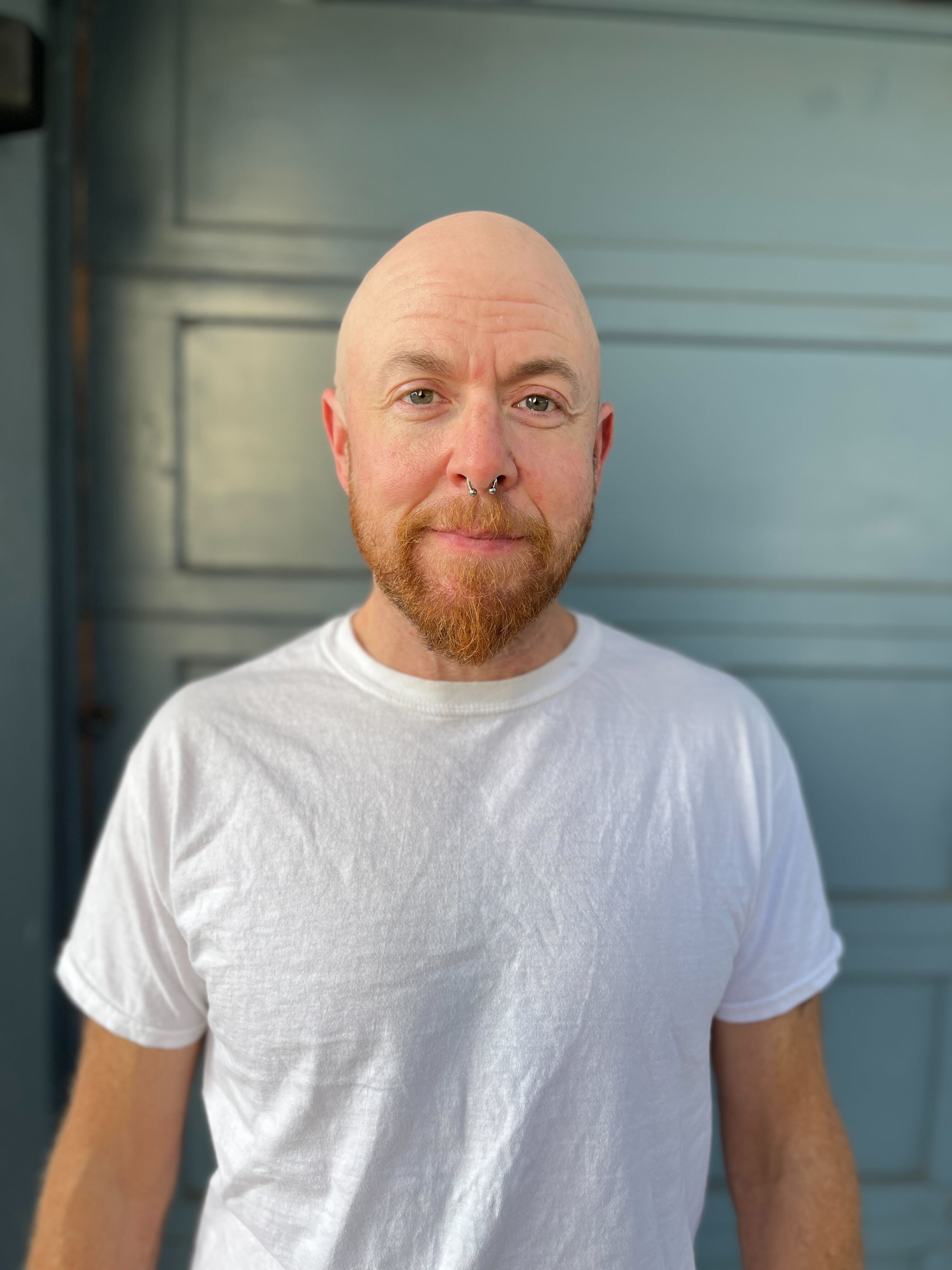 A bald man with a red beard and a septum piercing wears a plain white T-shirt, standing in front of a muted blue background.