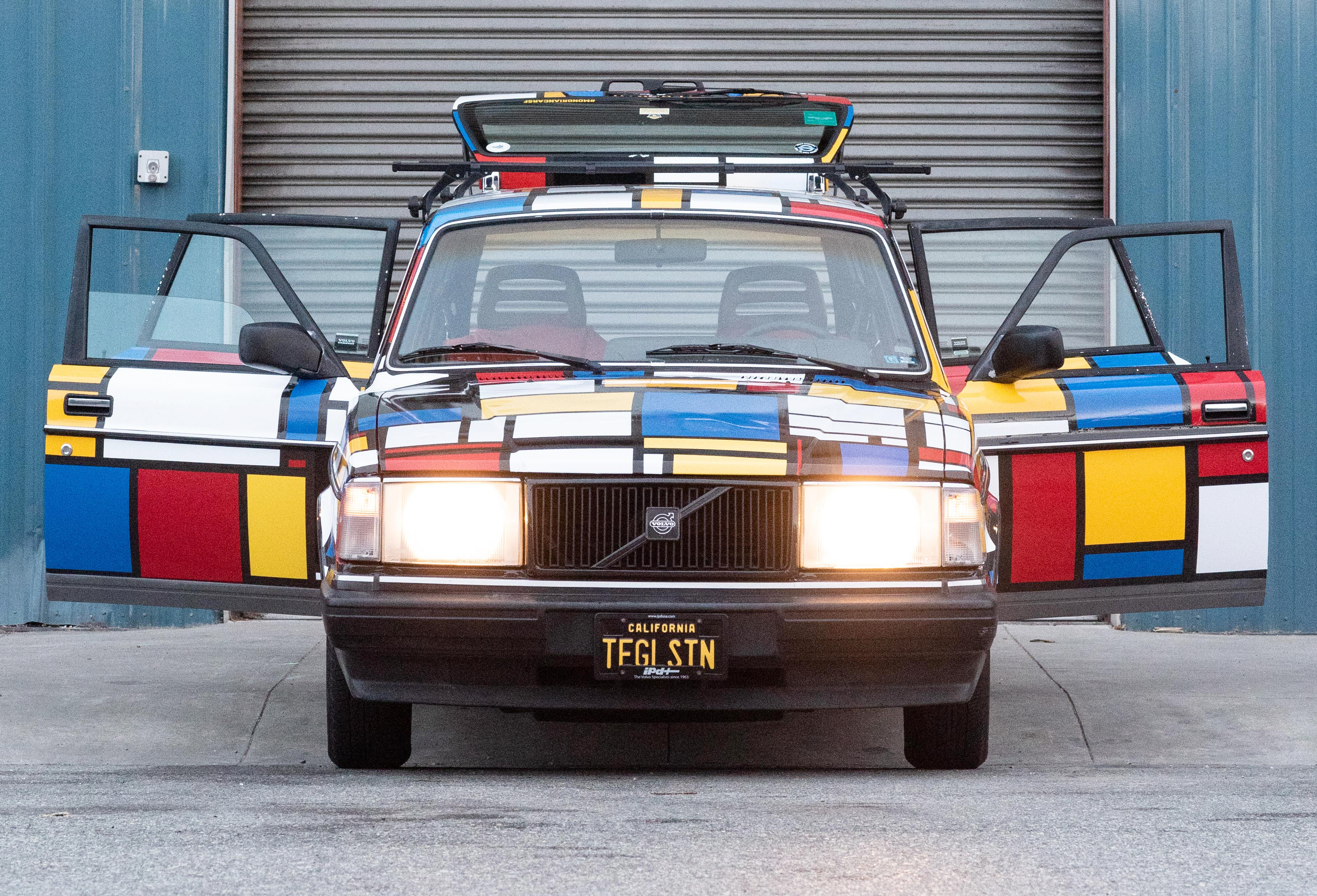 A car with Mondrian-inspired colorful geometric patterns has its doors open, revealing a license plate that reads &quot;TEGLSTN&quot; in front of a closed garage.