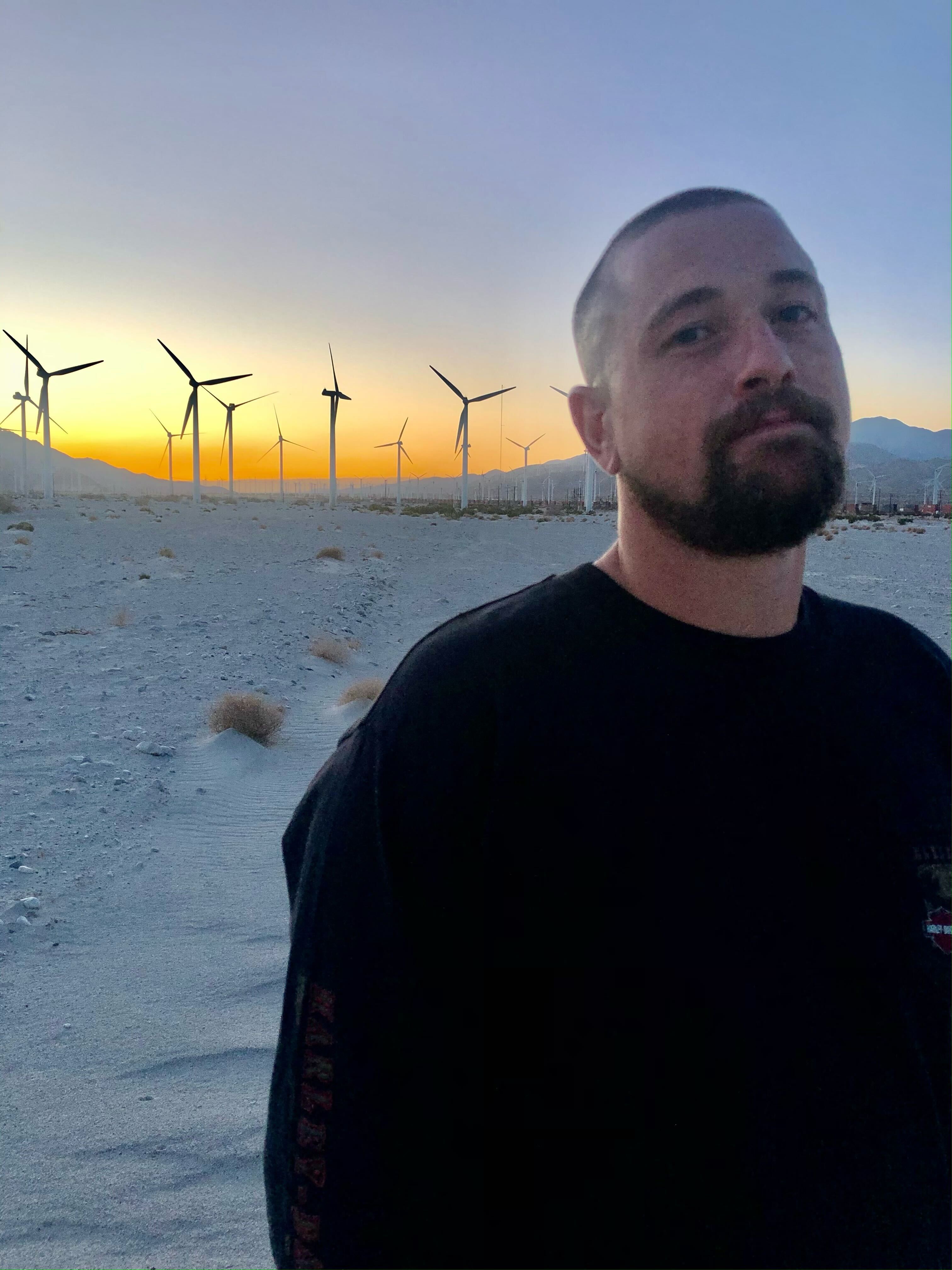 A person in a black shirt stands in the foreground, with wind turbines in a sandy desert and a colorful sunset sky in the background.