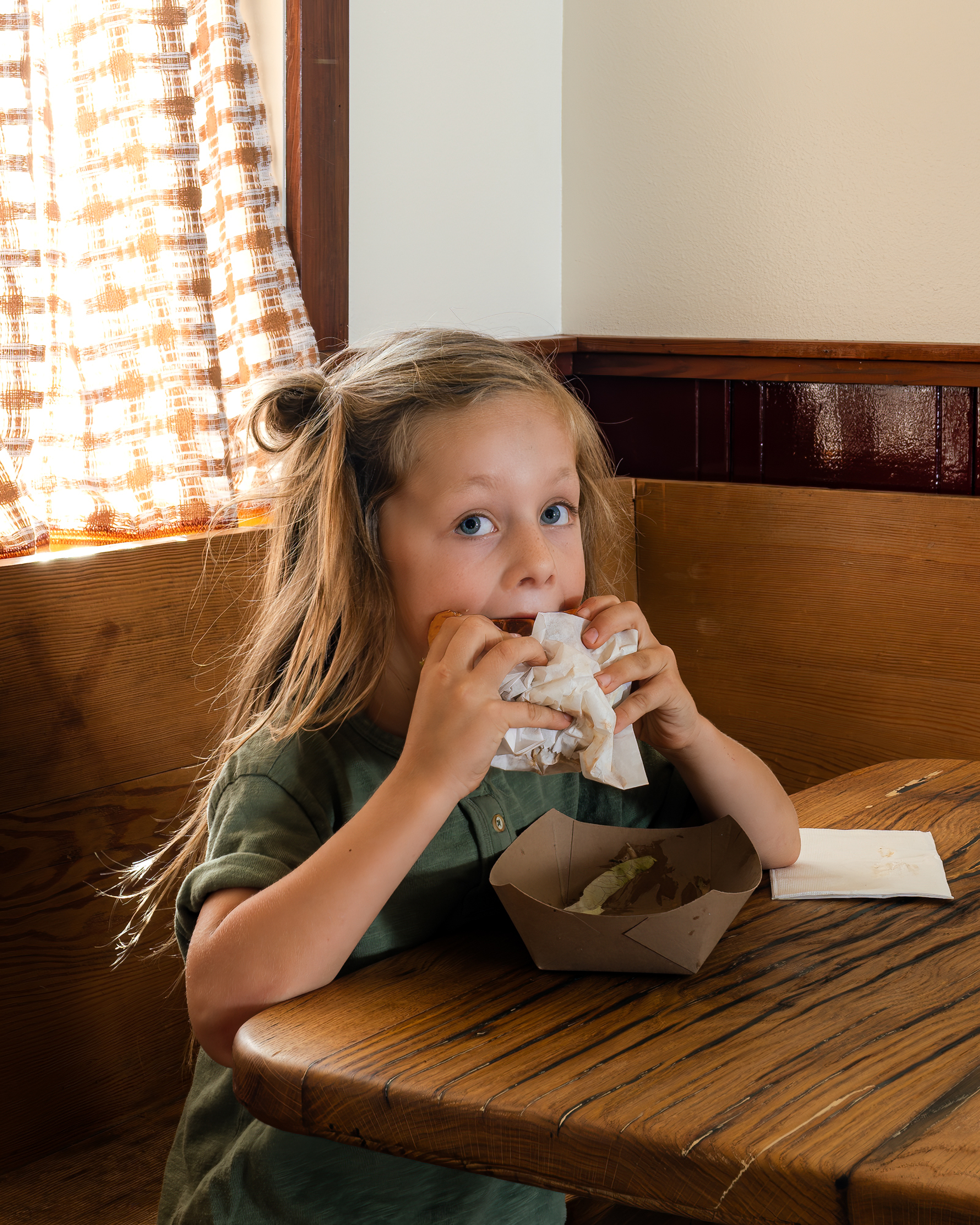 A child with long hair is sitting at a wooden table, enjoying a large sandwich. Sunlight filters through a plaid curtain, illuminating the cozy setting.