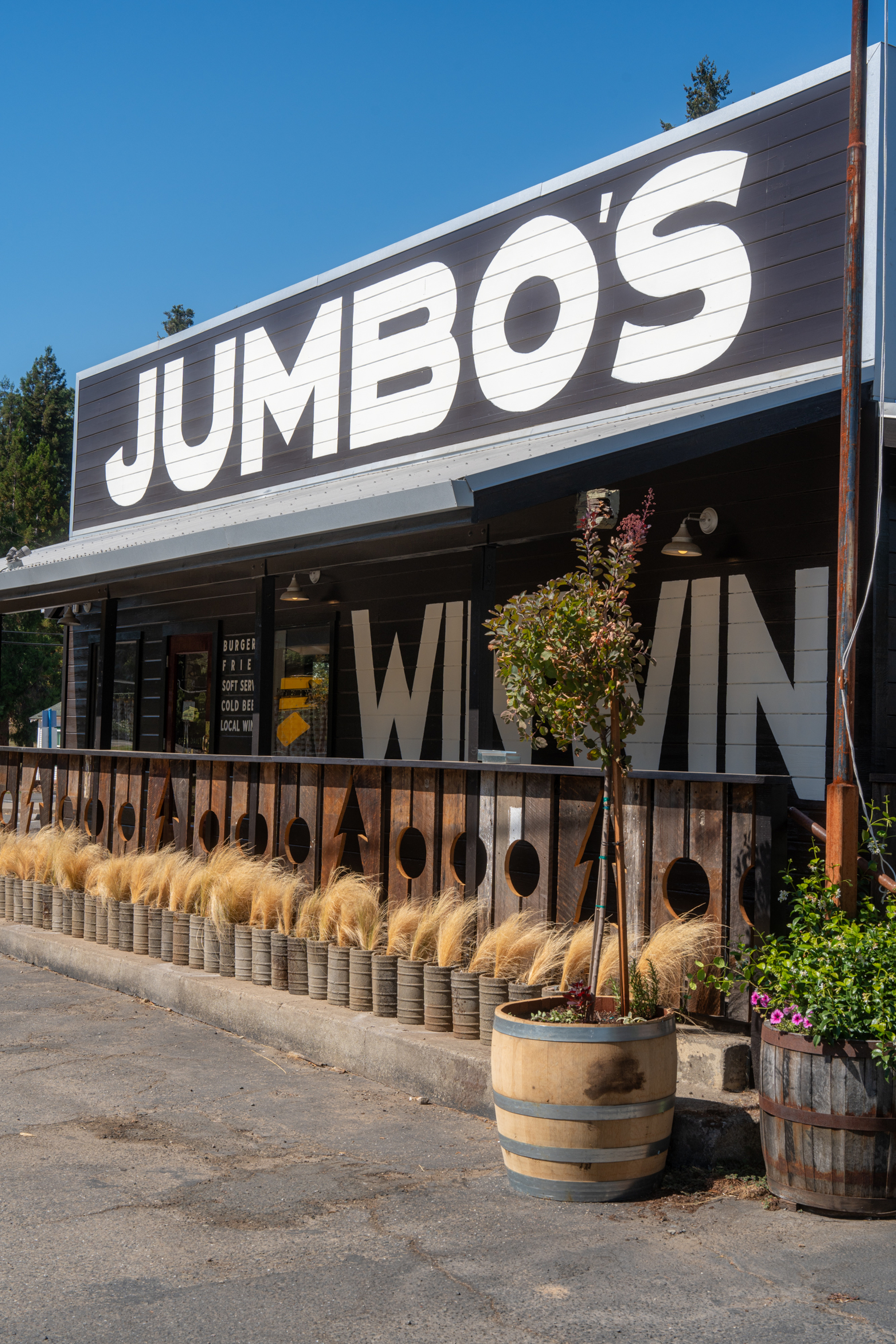 The image shows a roadside diner named &quot;JUMBO'S&quot; with a rustic wooden facade, surrounded by potted plants and a sign offering burgers, fries, and drinks.