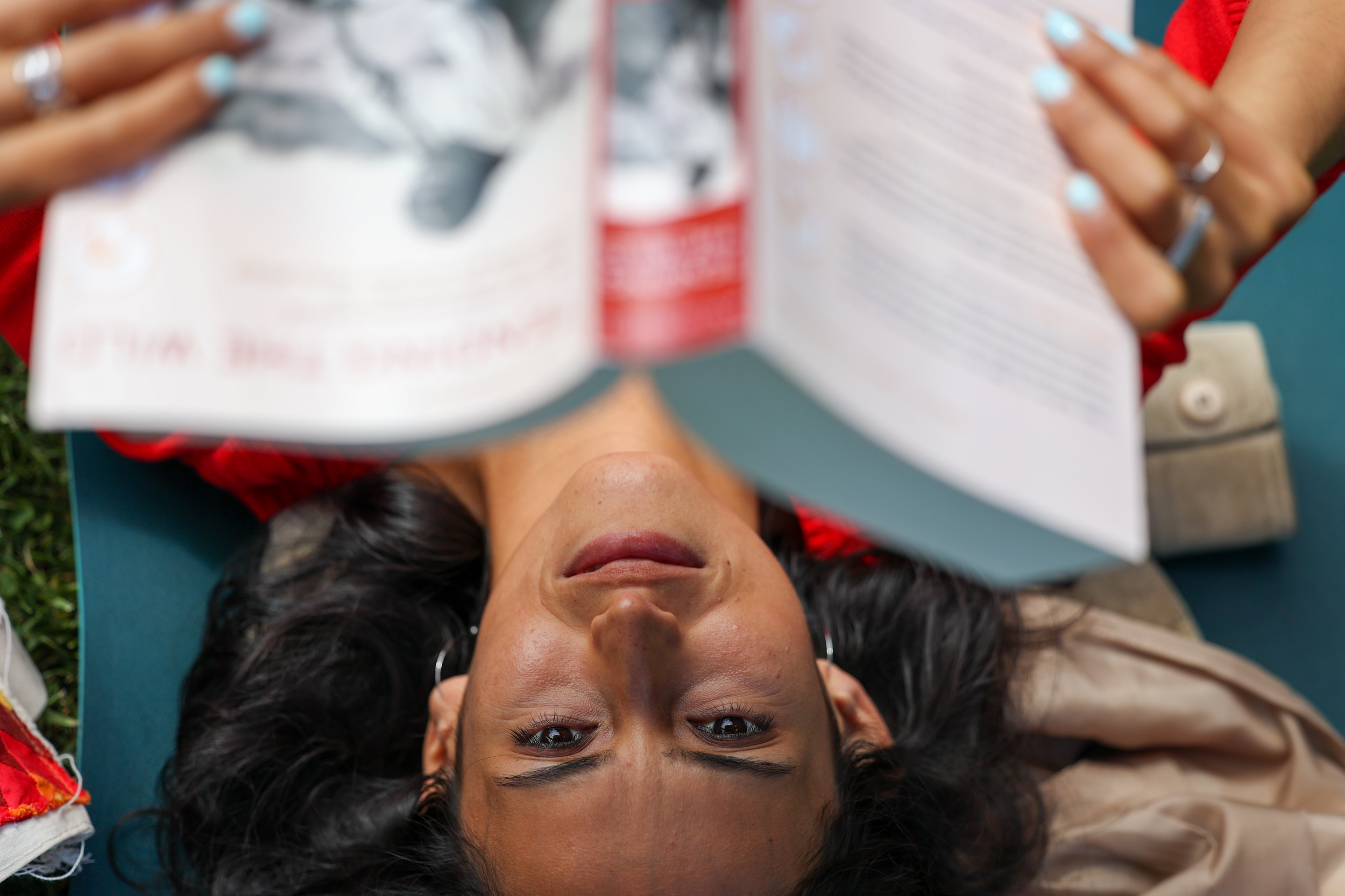 A woman with long dark hair is lying on her back, holding a book above her face. She appears relaxed, with a focused expression and painted nails.